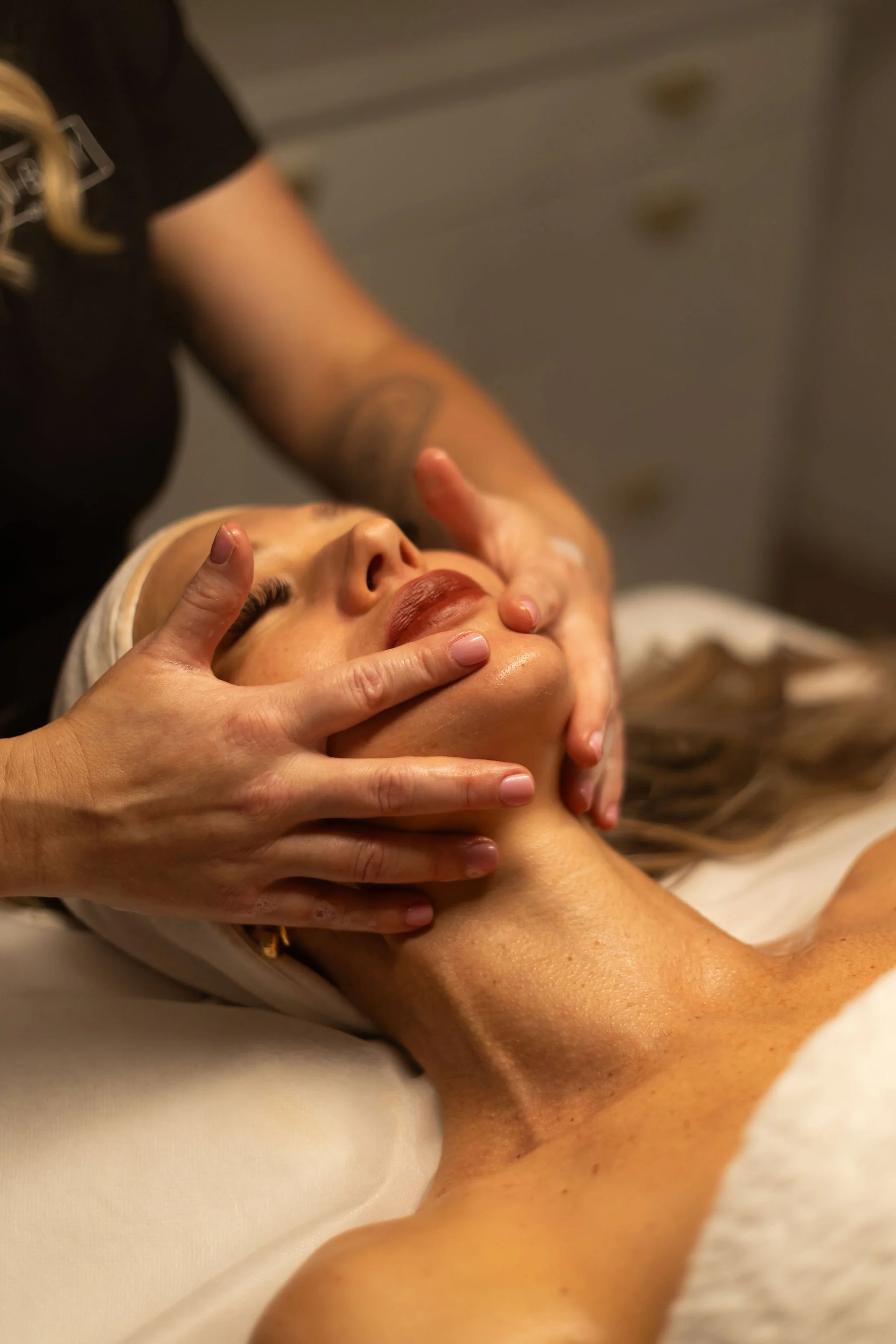 A woman receiving a facial treatment, lying down with her eyes closed, while a professional gently massages her face and neck.