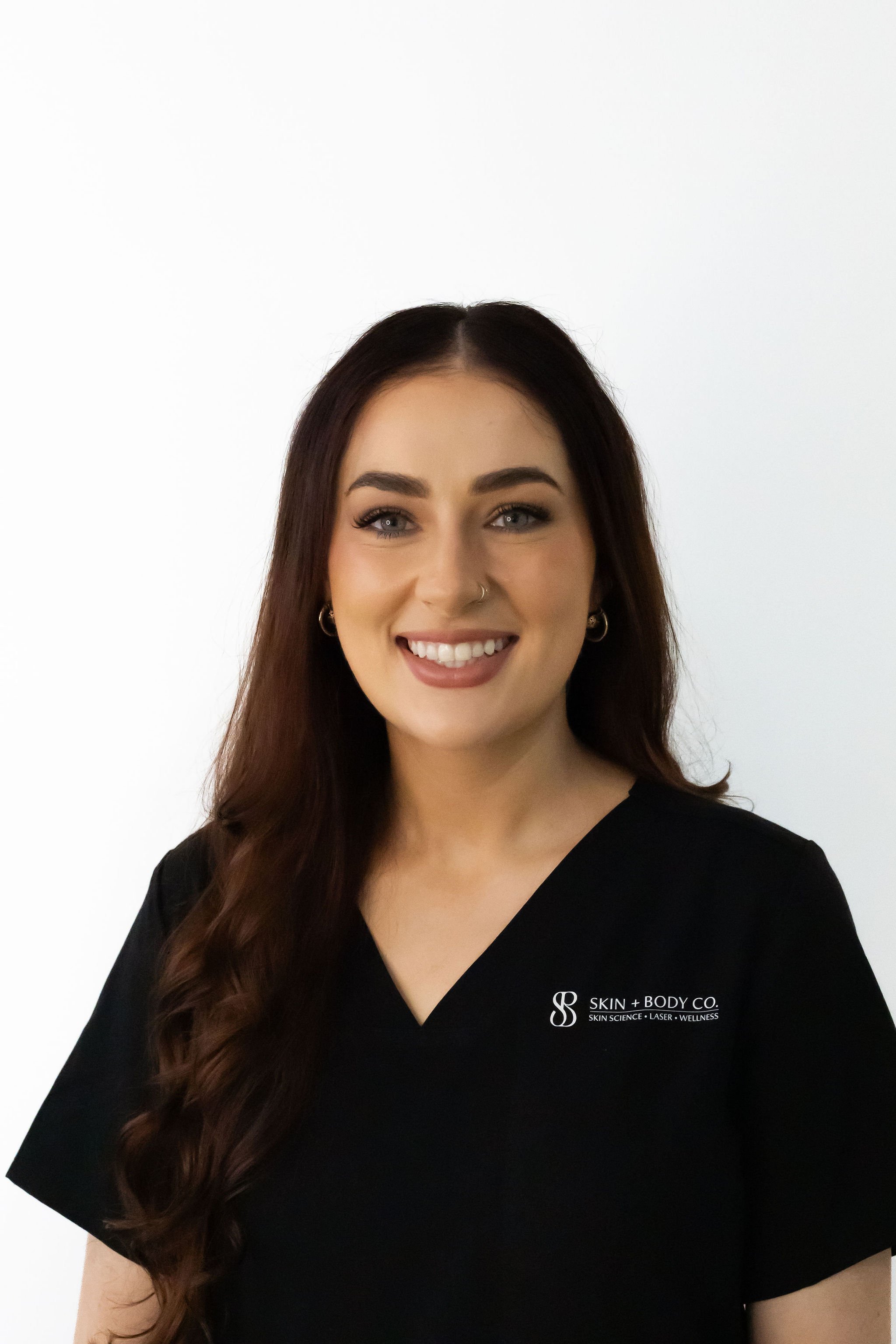 Close-up of a smiling woman with long, wavy brown hair, wearing black scrubs with the logo 'Skin + Body Co.' on the left chest, standing against a plain white background.
