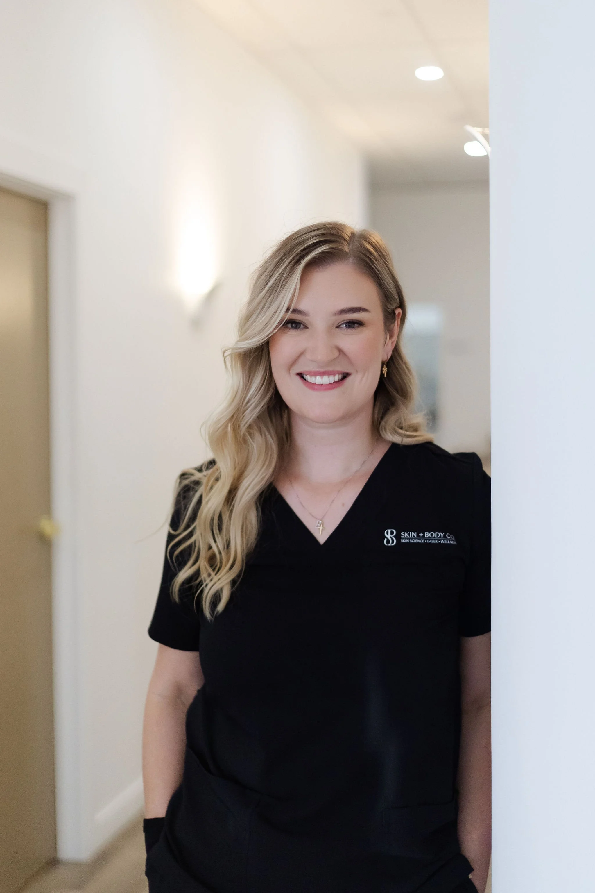 A woman with long blonde wavy hair smiling, wearing a black medical uniform with a logo, standing in a well-lit corridor.
