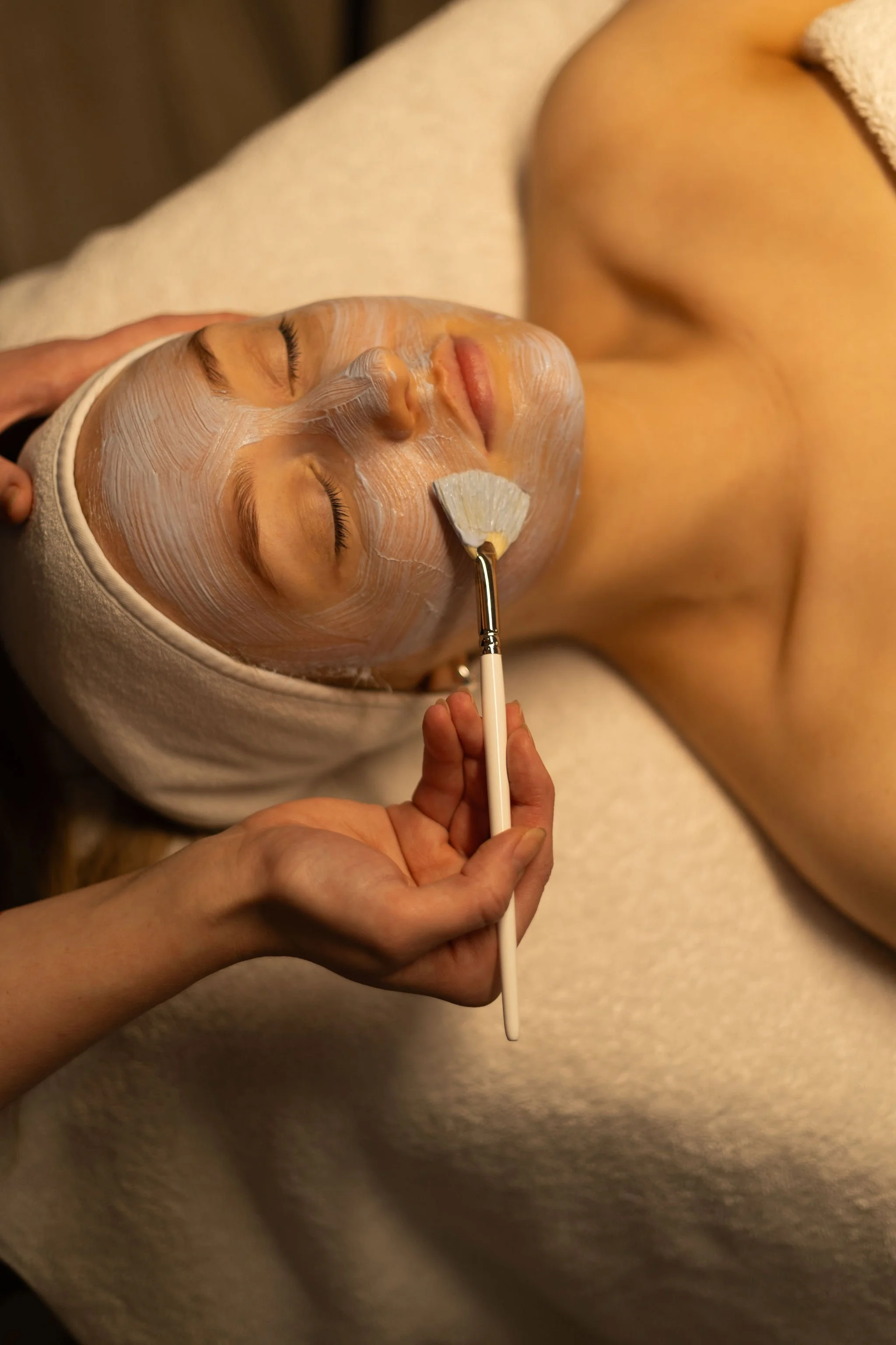 A woman receives a facial treatment with a white facial mask and a fan-shaped applicator, lying on a bed with a beige towel over her head.