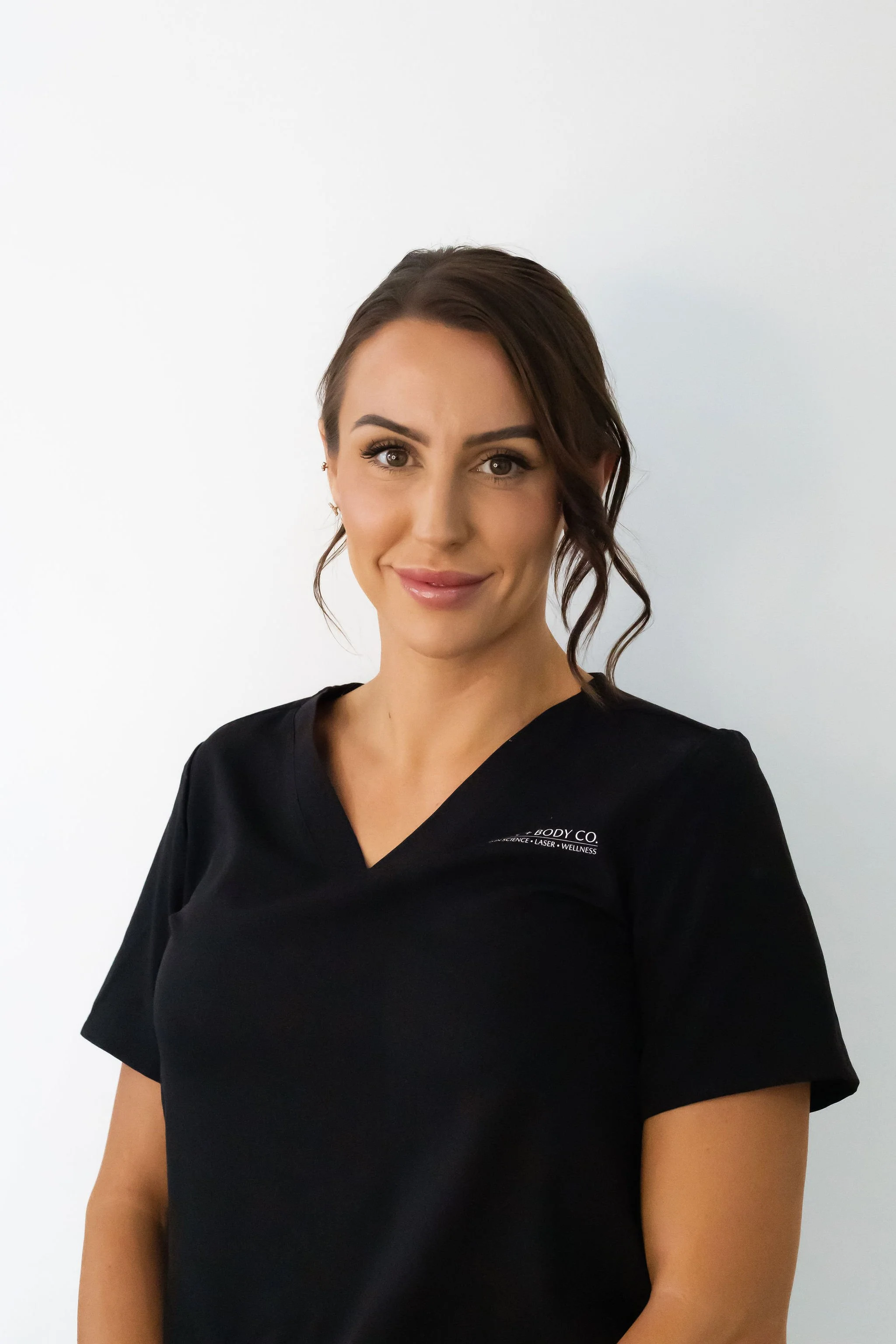 A smiling woman with brown hair styled in loose curls, wearing a black medical scrub top with a logo on the chest, standing against a plain white background.