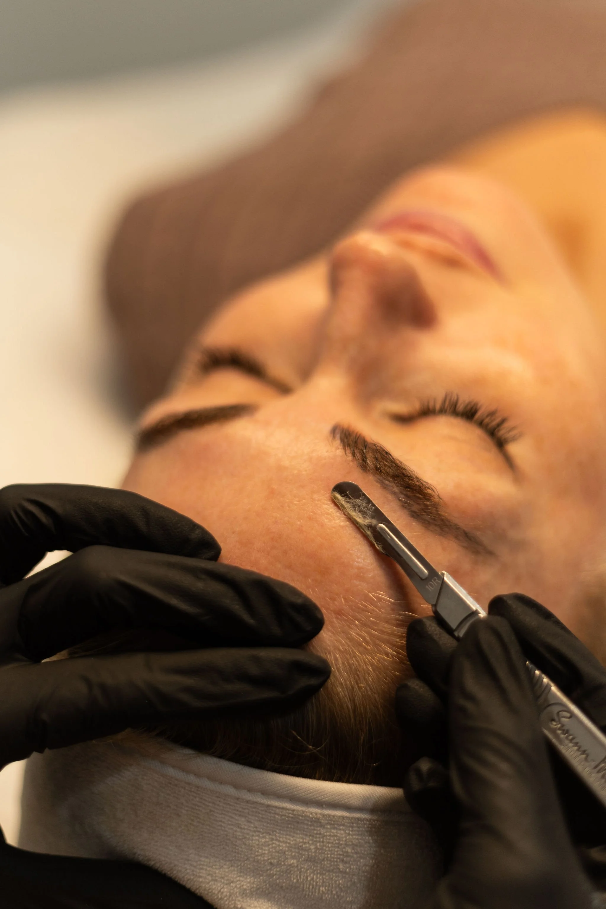 Close-up of a person's face with eyes closed, receiving an eye treatment or cosmetic procedure with a small tool, while wearing black gloves.