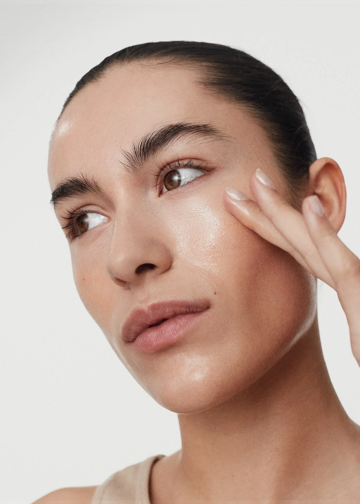Close-up of a woman with clear skin and dark hair pulled back, gently touching her face with her fingers.
