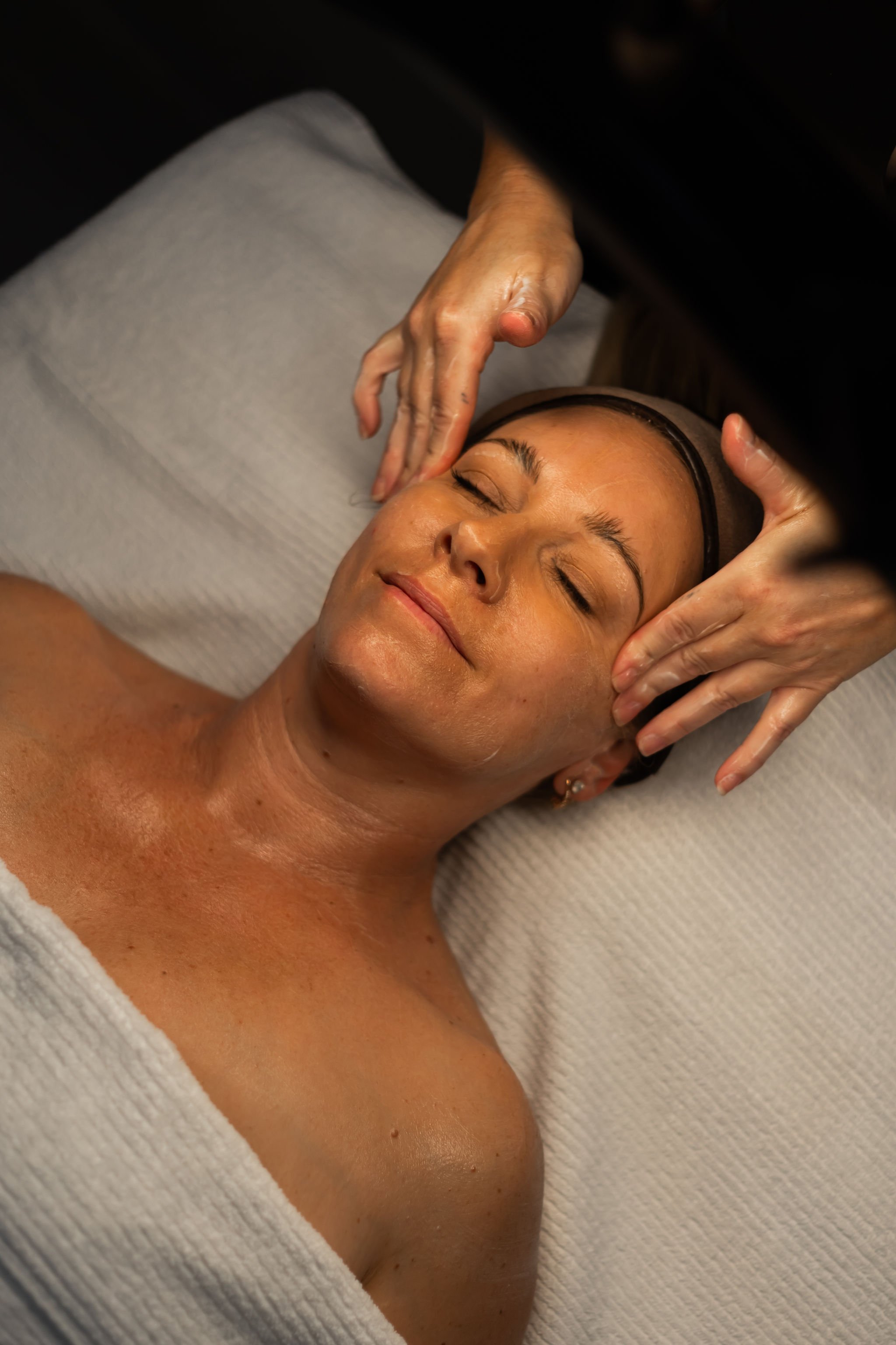 A woman is lying on a massage table with her eyes closed, receiving a facial treatment from an esthetician. The esthetician's hands are near the woman's face, applying a skincare product.
