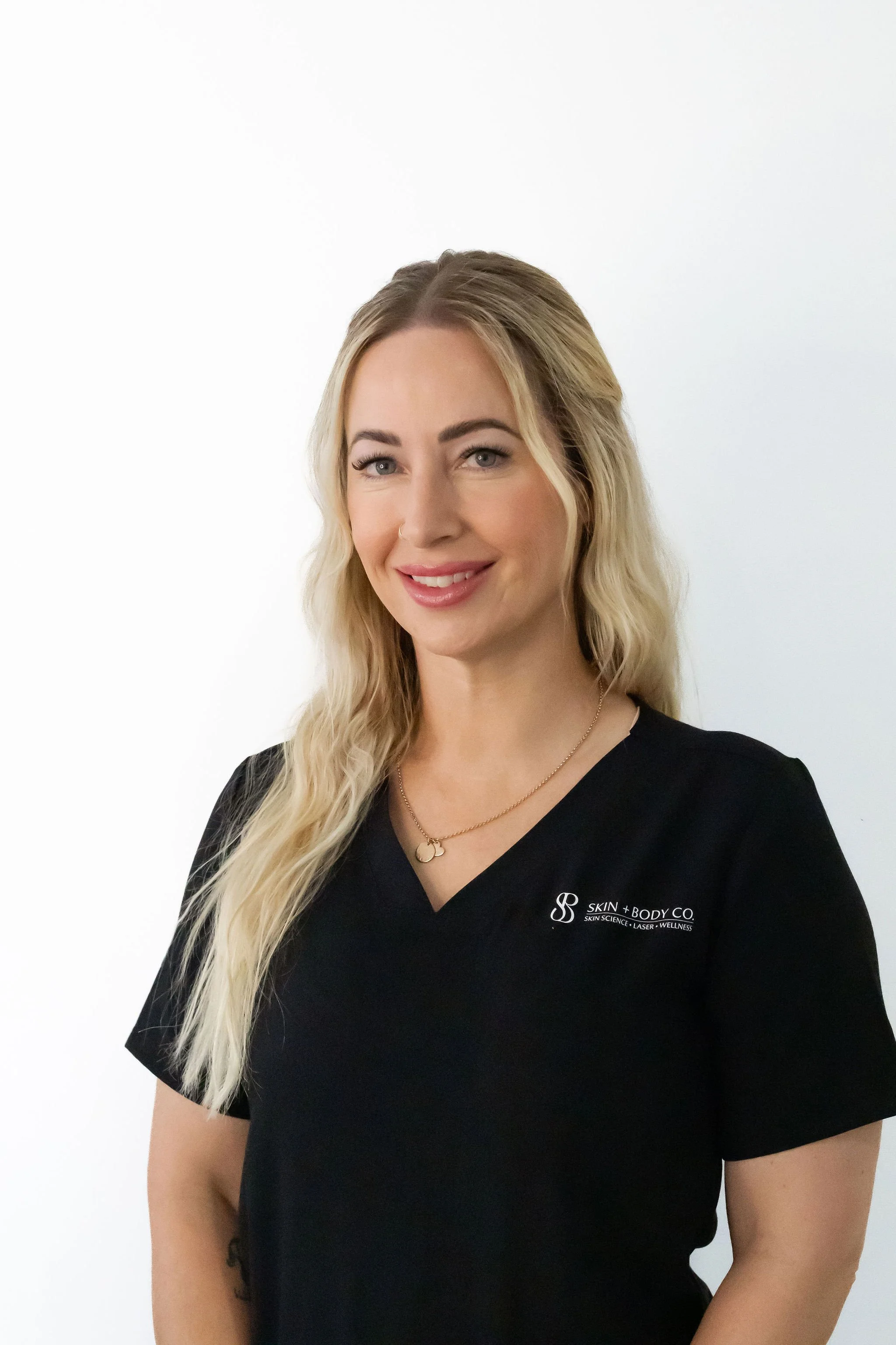 A woman with long, wavy blonde hair, light skin, and blue eyes smiling slightly, wearing a black T-shirt with the logo of Skin & Body Co. visible on the left side, standing against a white background.