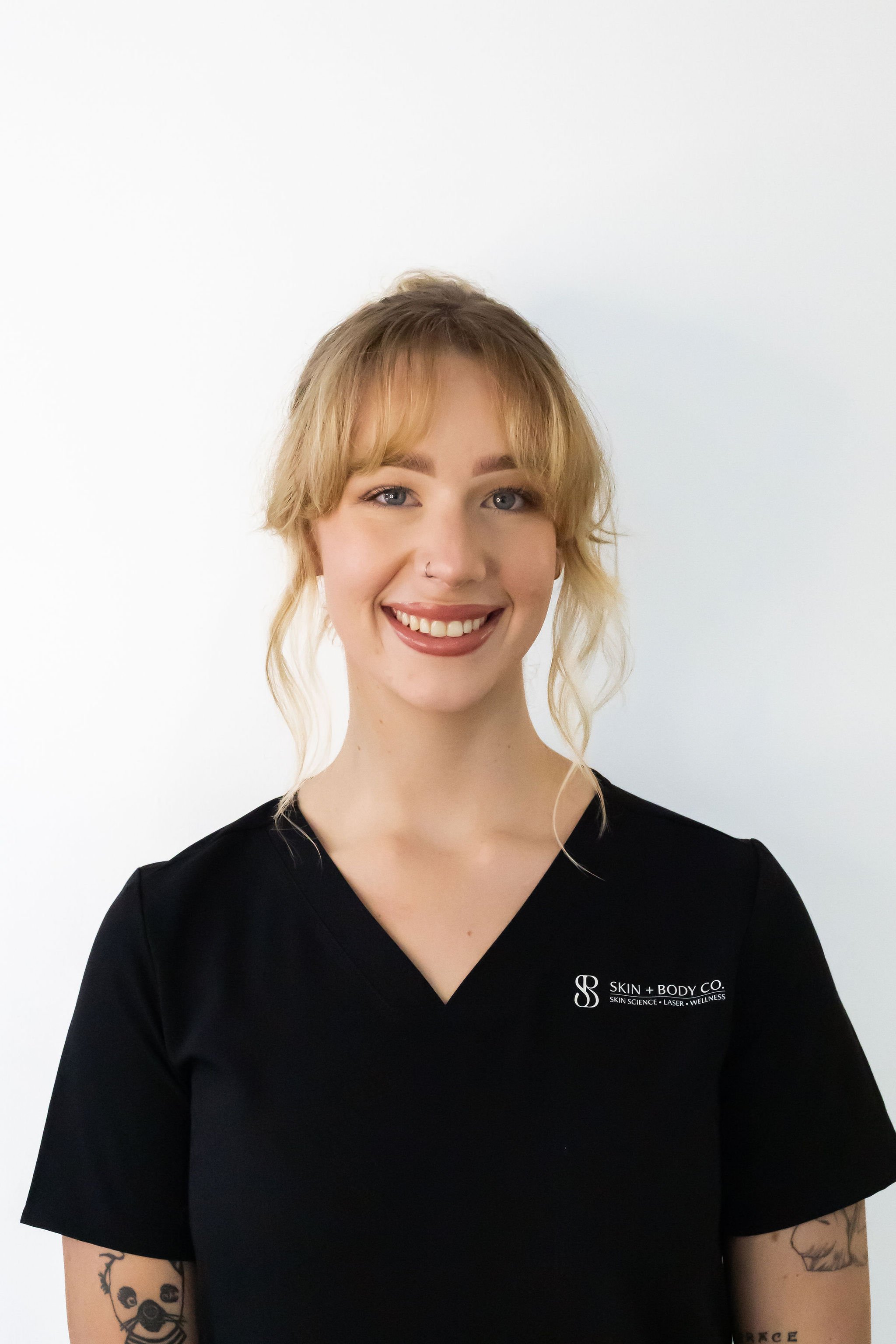 A smiling woman with blonde hair and tattoos on her arms, wearing a black medical uniform with the logo 'Skin + Body Co.' on it, standing against a white background.