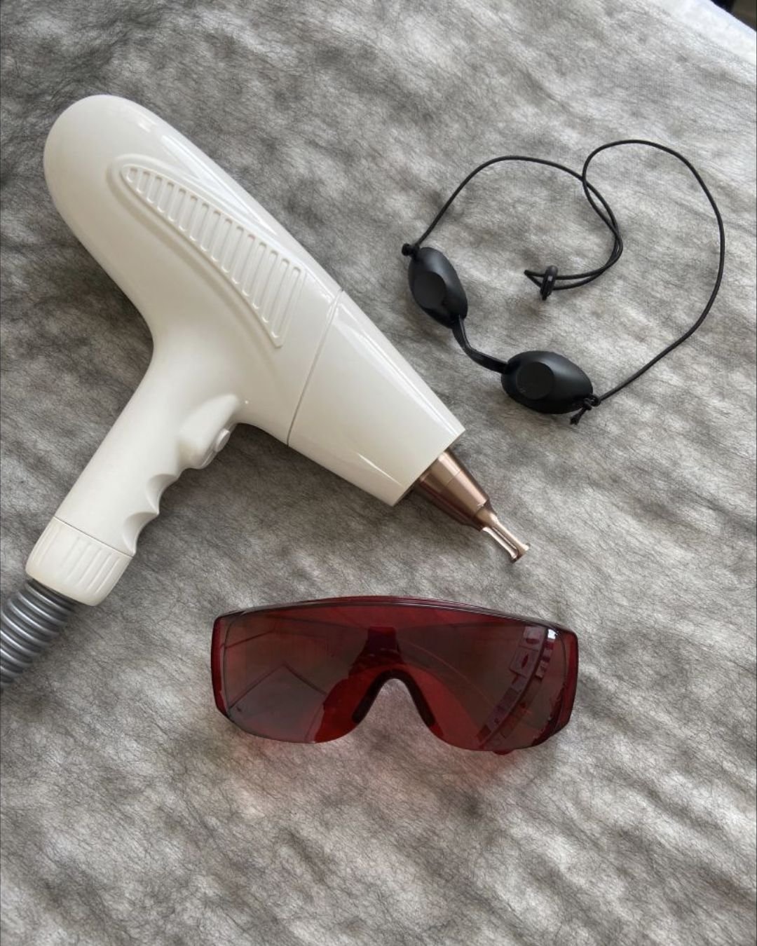 A white handheld device with a metallic tip, a black headset with an adjustable strap, and a pair of red protective glasses on a gray textured surface.