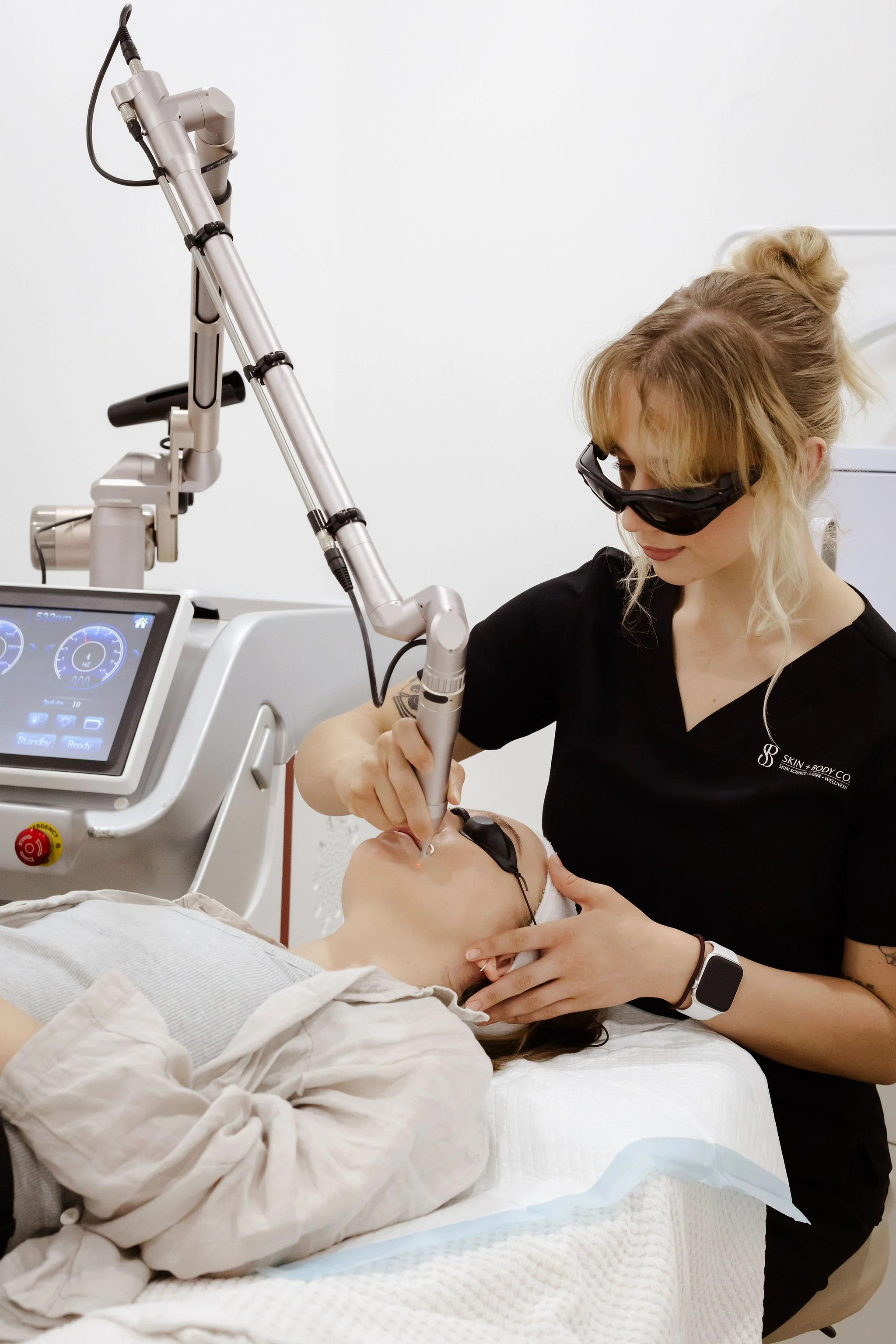 A woman performing a facial treatment on a patient in a medical setting. The patient is lying down with a facial mask, and the woman is using specialized equipment while wearing protective glasses.
