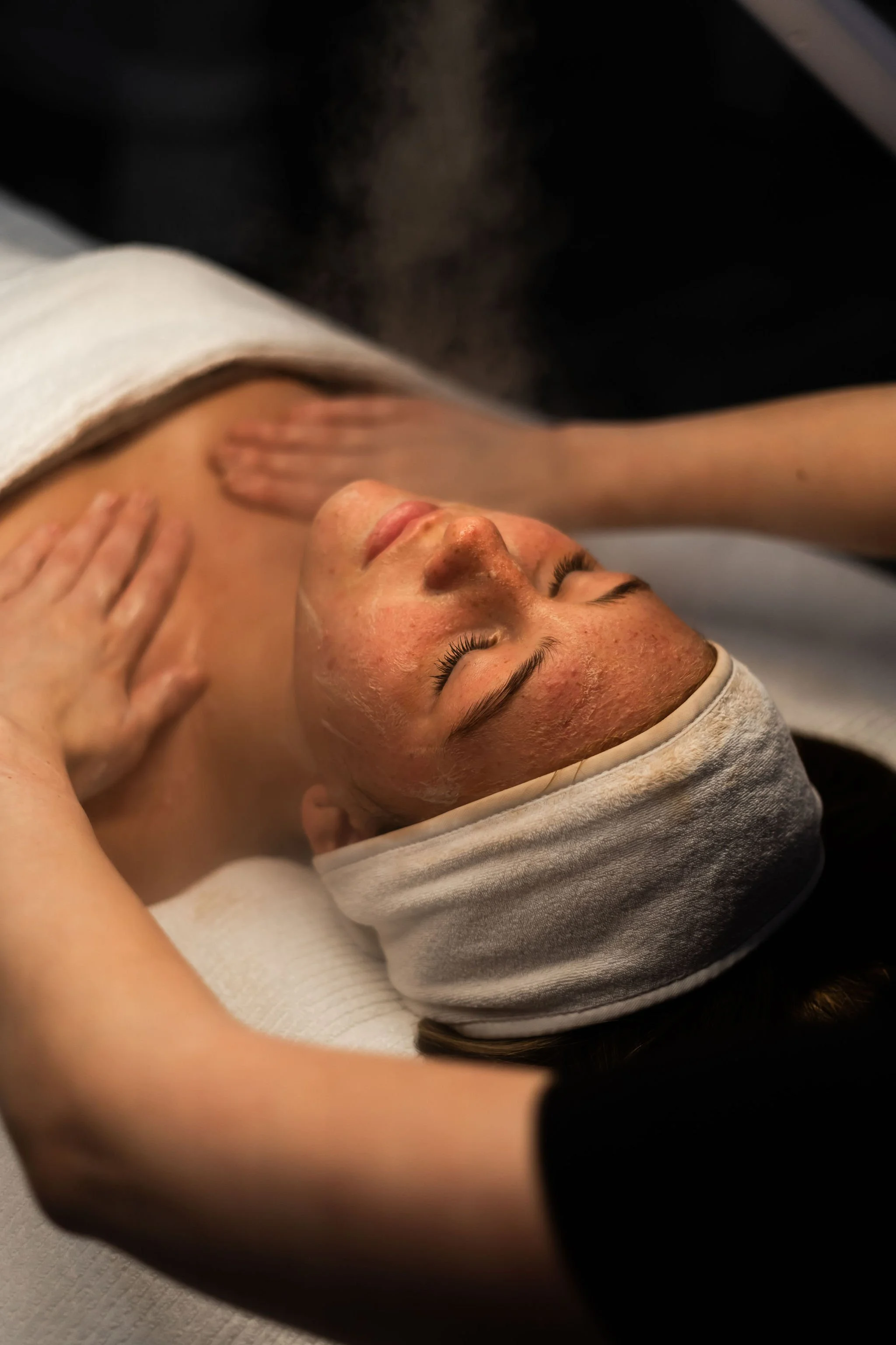 Person lying down during a facial treatment or skincare procedure, wearing a towel and cap, with facial steam or mist visible.