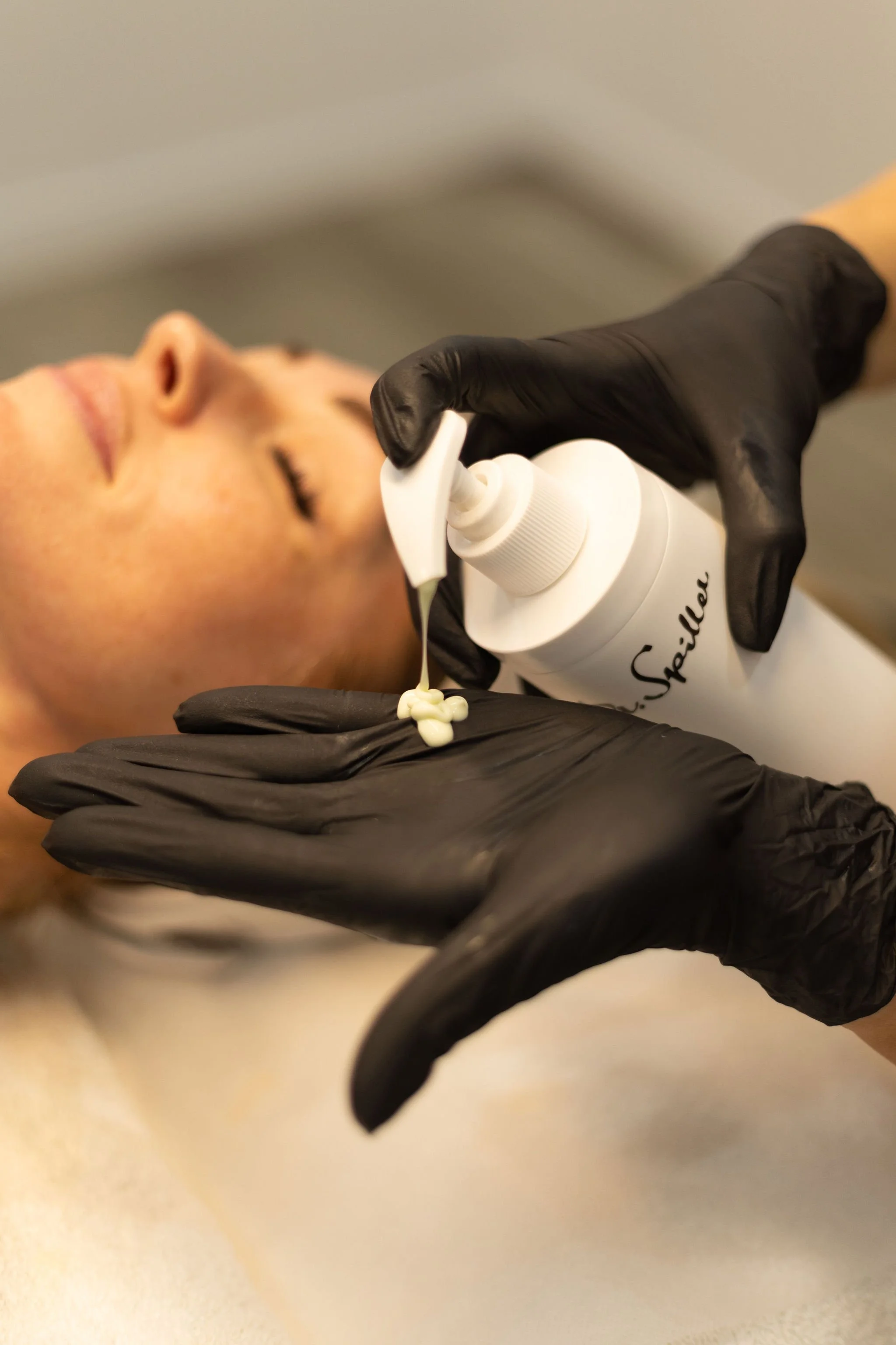 A person lying down with their eyes closed, receiving a facial treatment from a professional at Skin + Body Co wearing black gloves, applying a skincare product from a bottle.