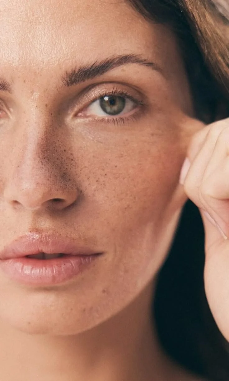 Close-up of a woman's face showing her eye, nose, and lips with natural makeup and freckles, with her hand touching her cheek.
