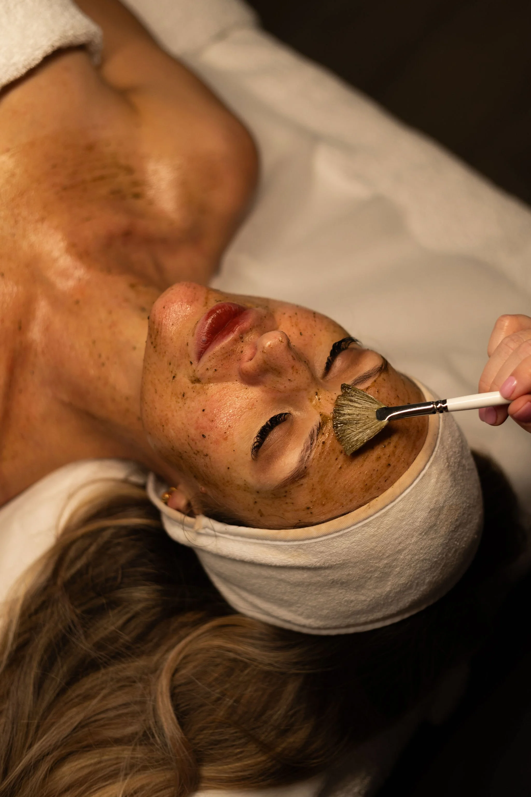 A woman receiving a facial treatment, with her face relaxed and eyes closed, lying on a treatment bed with a towel wrapped around her head, and a brush applying a facial mask.