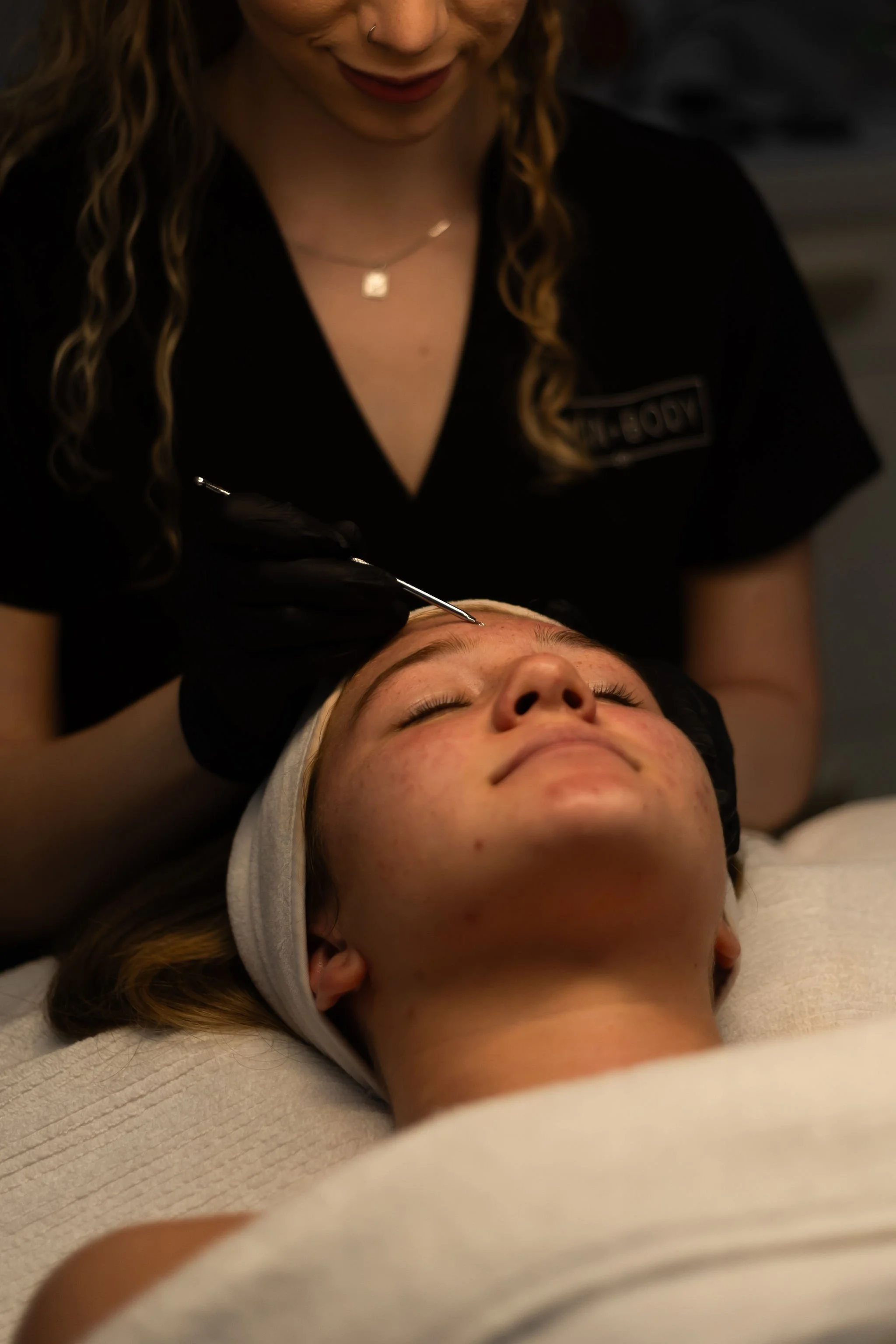 A woman with curly hair, wearing a black shirt with a logo, is performing a procedure on a person lying down with their eyes closed, wearing a hoodie and a headband. The woman is smiling slightly and holding a tool near the person's forehead.