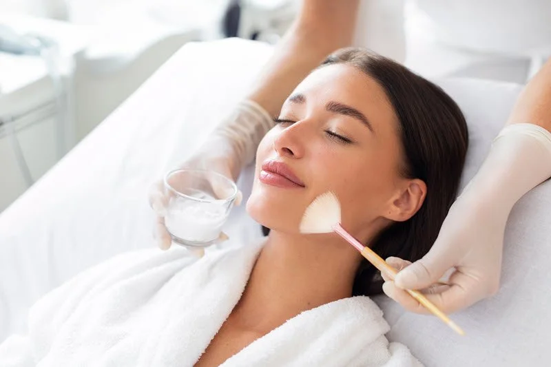 Woman getting a facial treatment, with a mask being applied to her face, wearing a white robe, and relaxing with closed eyes.