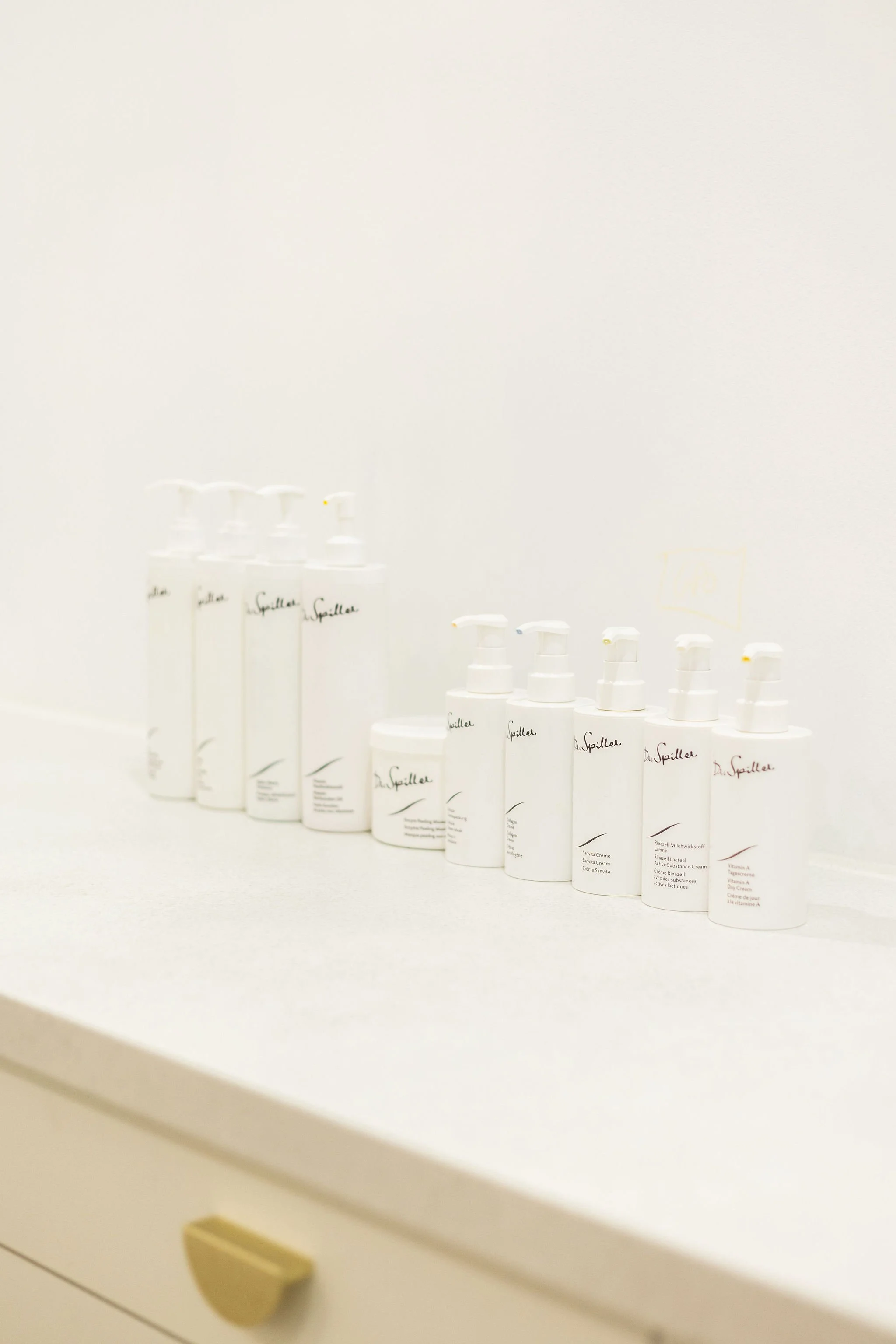 Several white bottles with pump dispensers lined up on a white surface in front of a white wall.