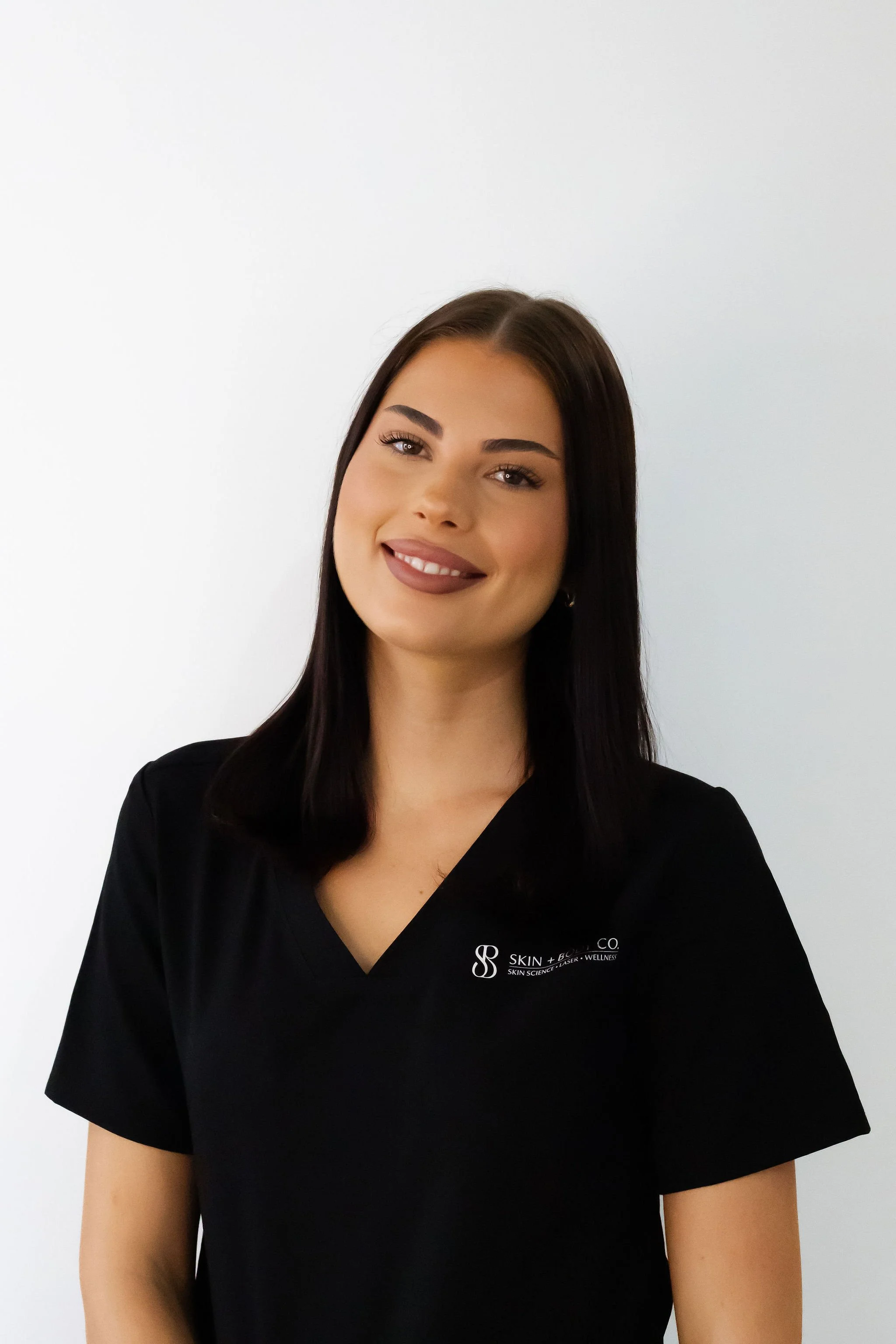 A young woman with long dark hair and a bright smile, wearing a black V-neck scrub top with a logo from Skin + B Co, standing against a plain white wall.