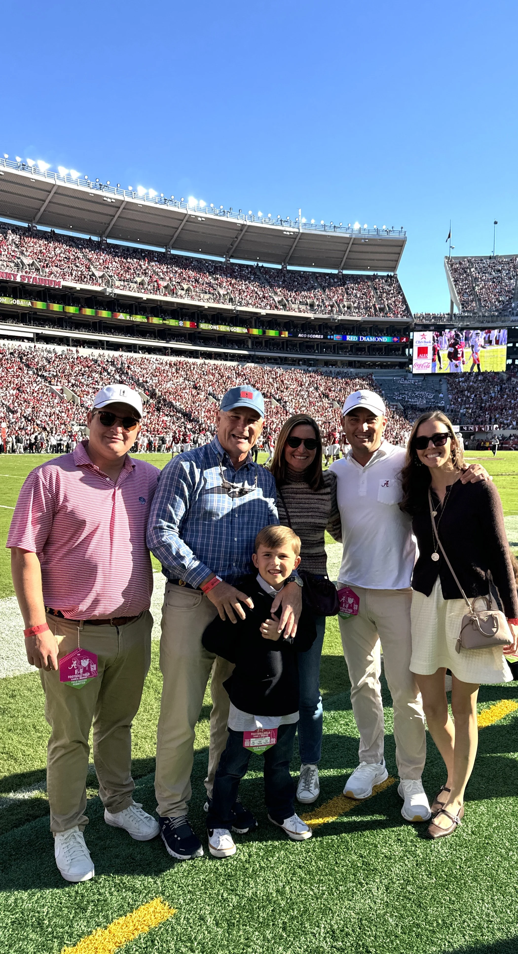 Six people, including a young boy, standing on a football field in a stadium filled with spectators during daytime, with a large screen displaying game action in the background.