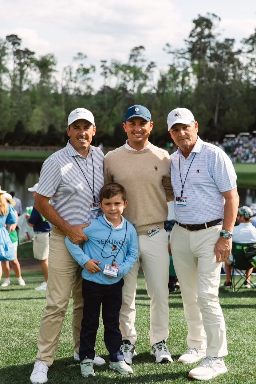Four people, three adults and one child, posing outdoors on a golf course near a water hazard. The adults are wearing golf attire and caps, and all are smiling at the camera.