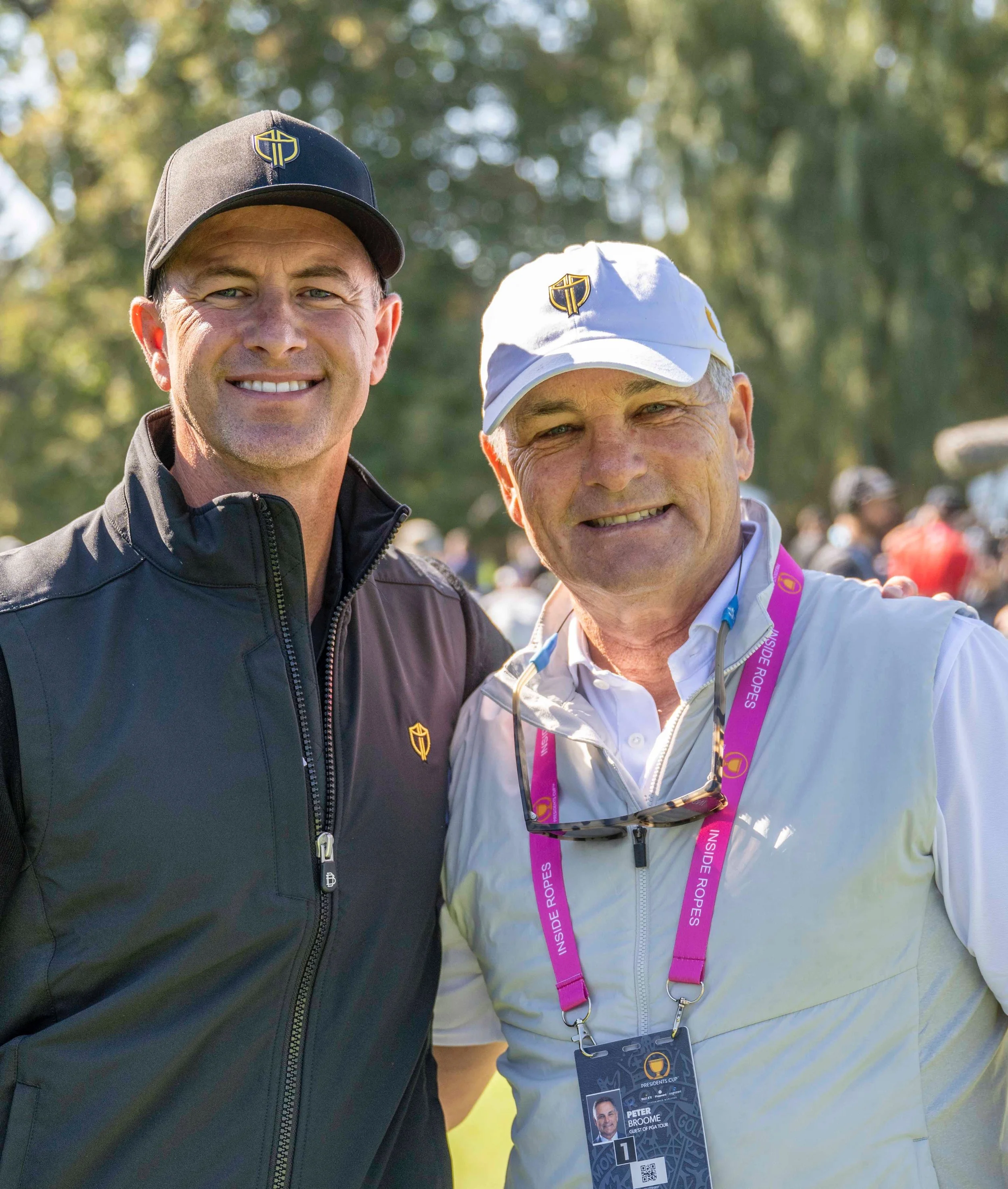 Two men standing outdoors, smiling, wearing golf attire and hats with logos.