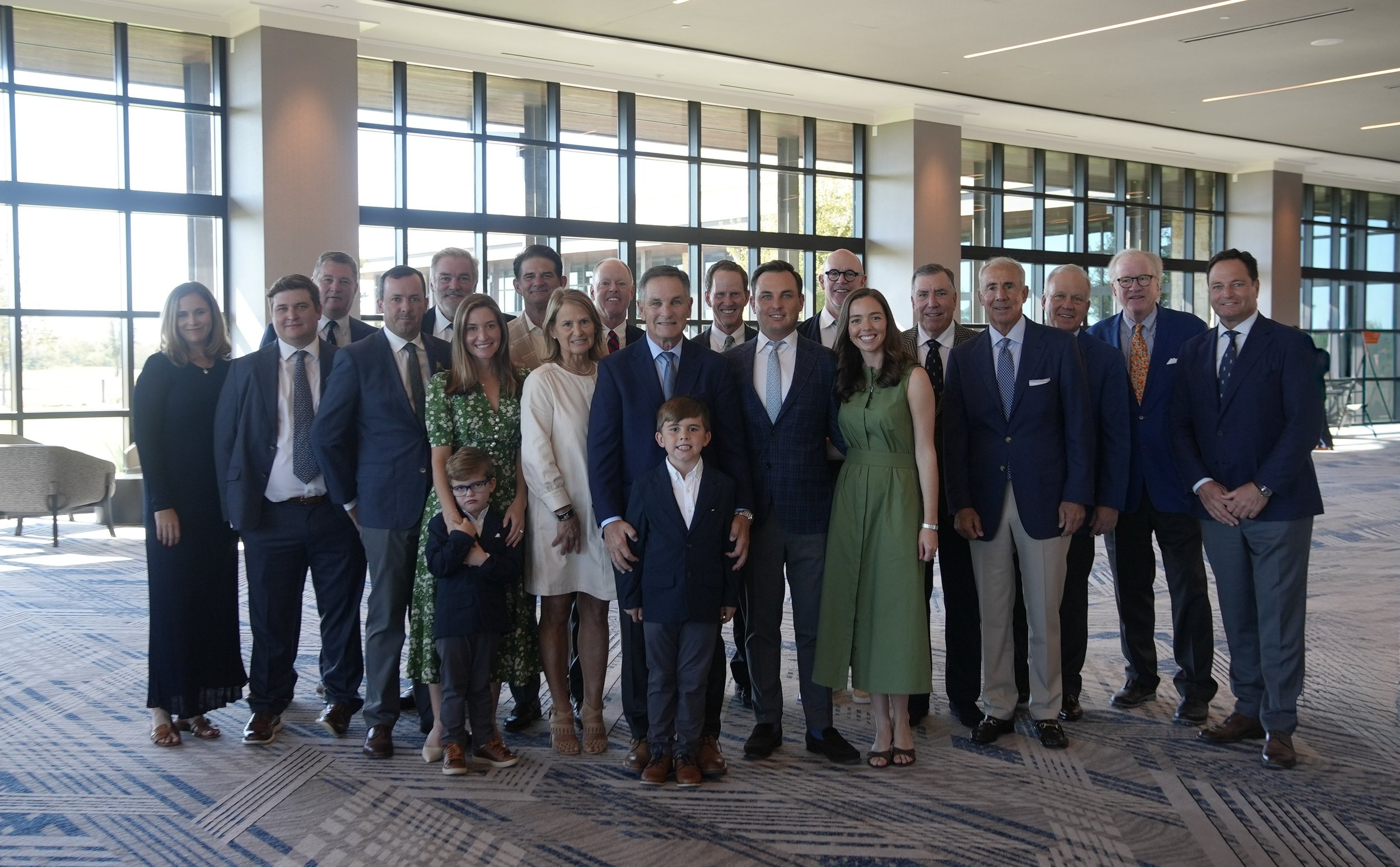 Group of professionally dressed people standing in a spacious, modern building with large windows and natural lighting.