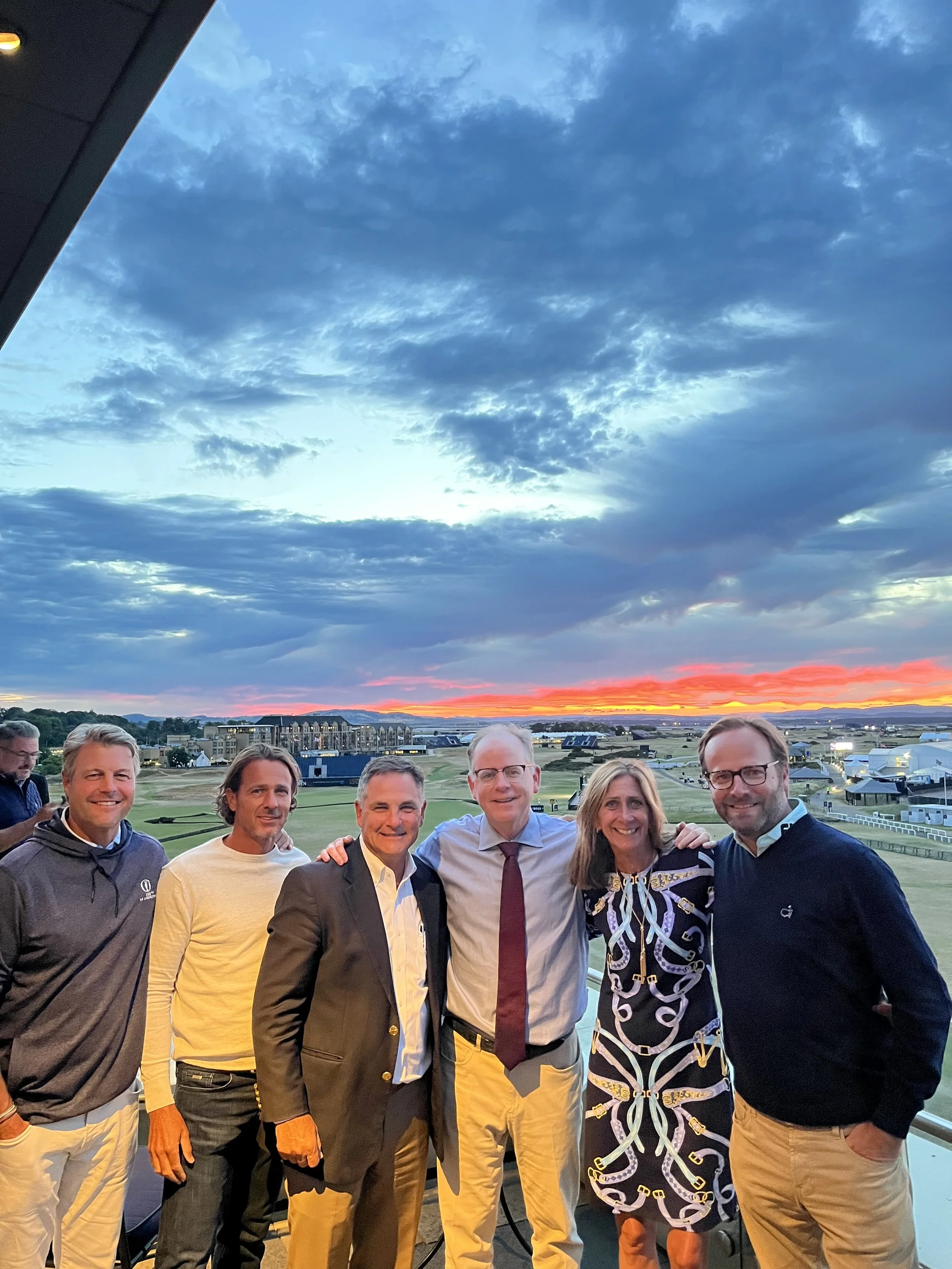 Group of six people standing on a balcony at sunset, smiling, with a grassy field and a cloudy sky in the background.