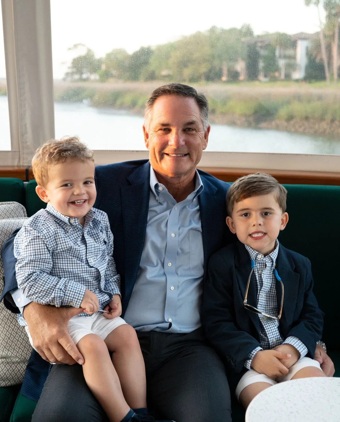 A smiling man sitting with two young boys in front of a window with a view of a river and trees. The boys are dressed in checkered shirts and blazers.