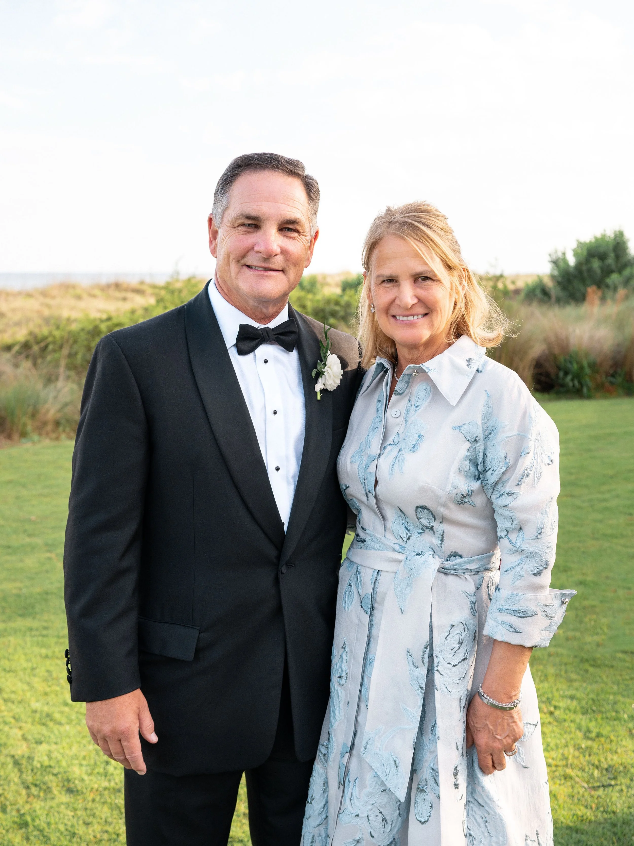 A man in a tuxedo and a woman in a light-colored dress stand outdoors on a grassy area, smiling at the camera.
