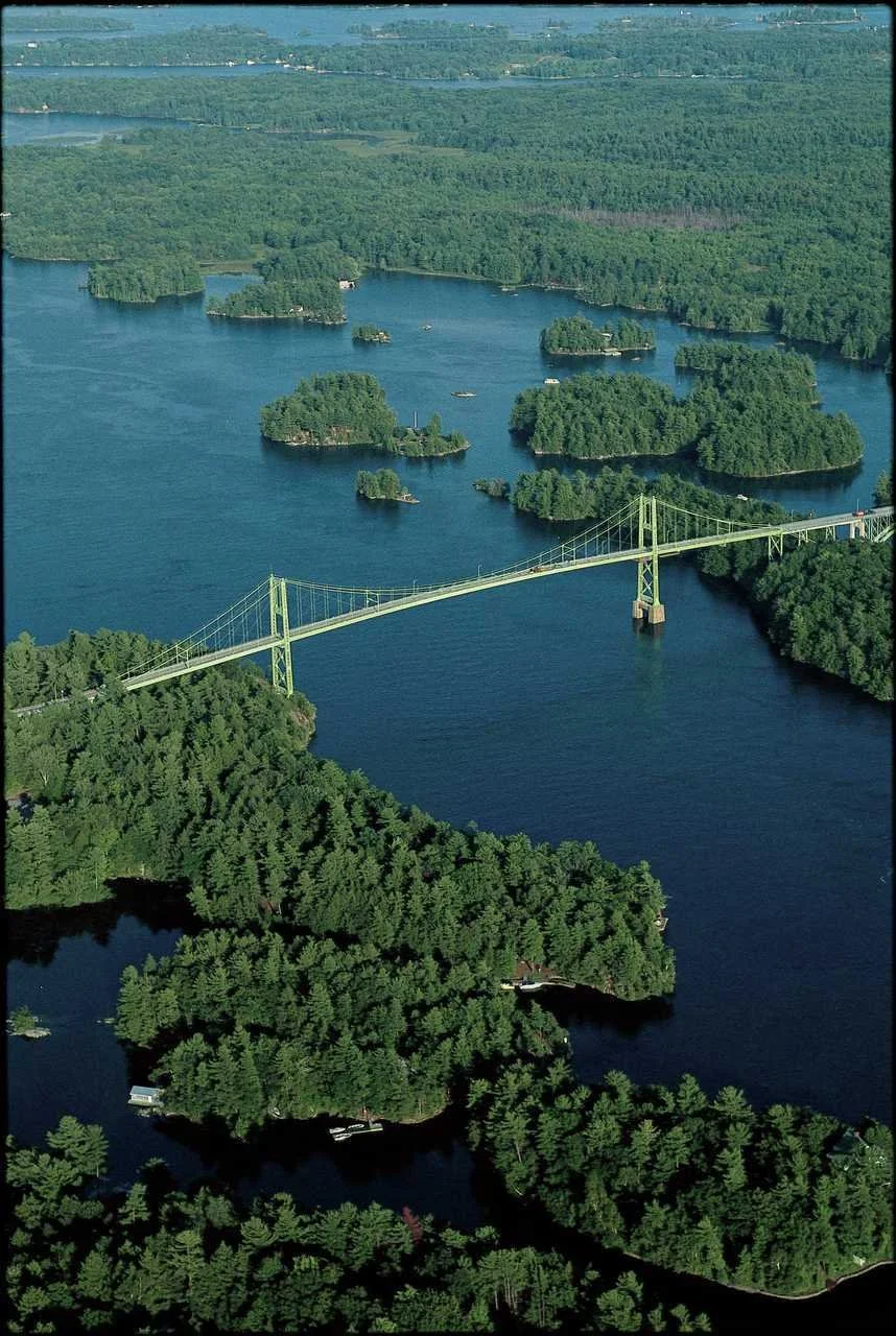 An aerial view of a large body of water with numerous small islands covered in dense green trees, and a long bridge stretching across the water connecting the landmasses. The scene is lush and green with a landscape of lakes, forests, and the bridge.
