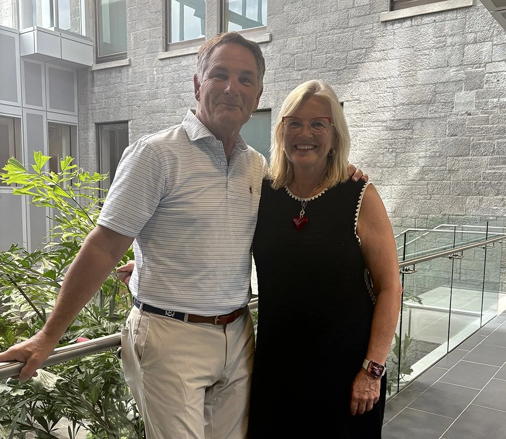 A smiling man and woman standing together inside a building with large windows and gray brick walls, greenery on the left, and glass railing on the right.