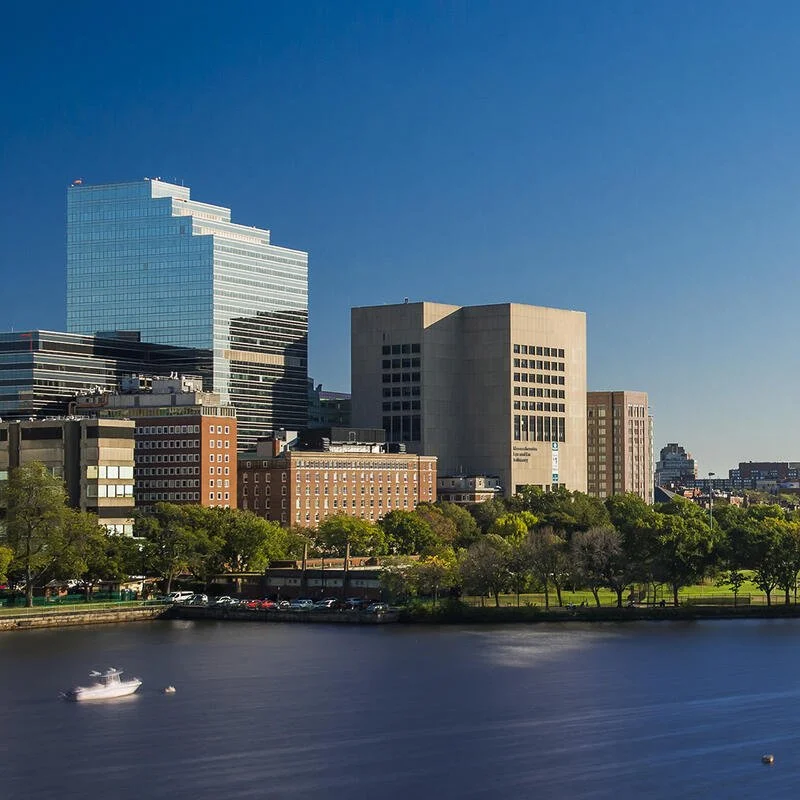 City skyline with modern skyscrapers beside a river, with a boat on the water and green trees along the riverbank.