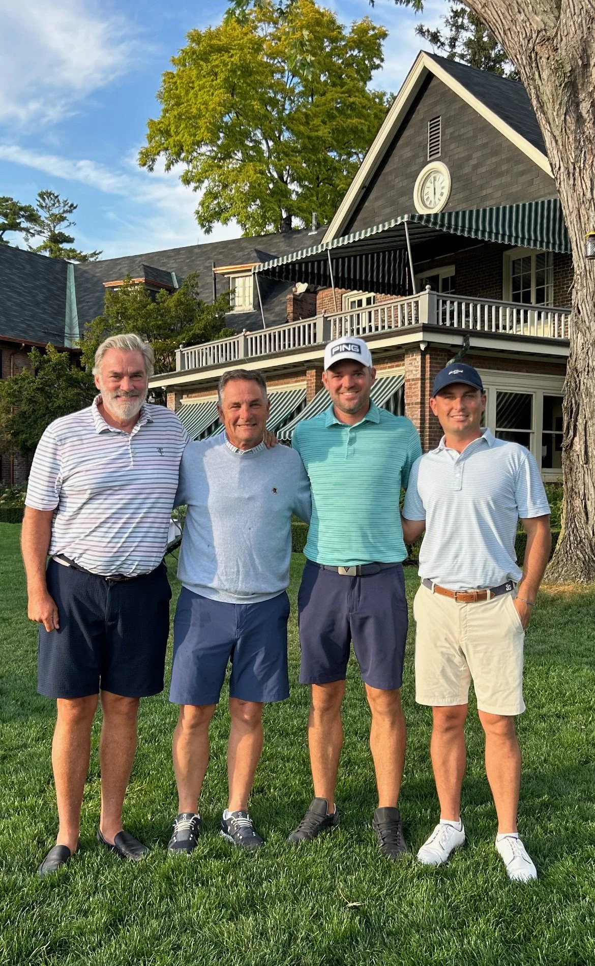 Four men standing on a grassy lawn in front of a large house, smiling at the camera. The men are dressed in golf attire, with the two on the right wearing golf hats. The house is brick and wood, with a second-floor balcony, striped awnings, and a large tree nearby.