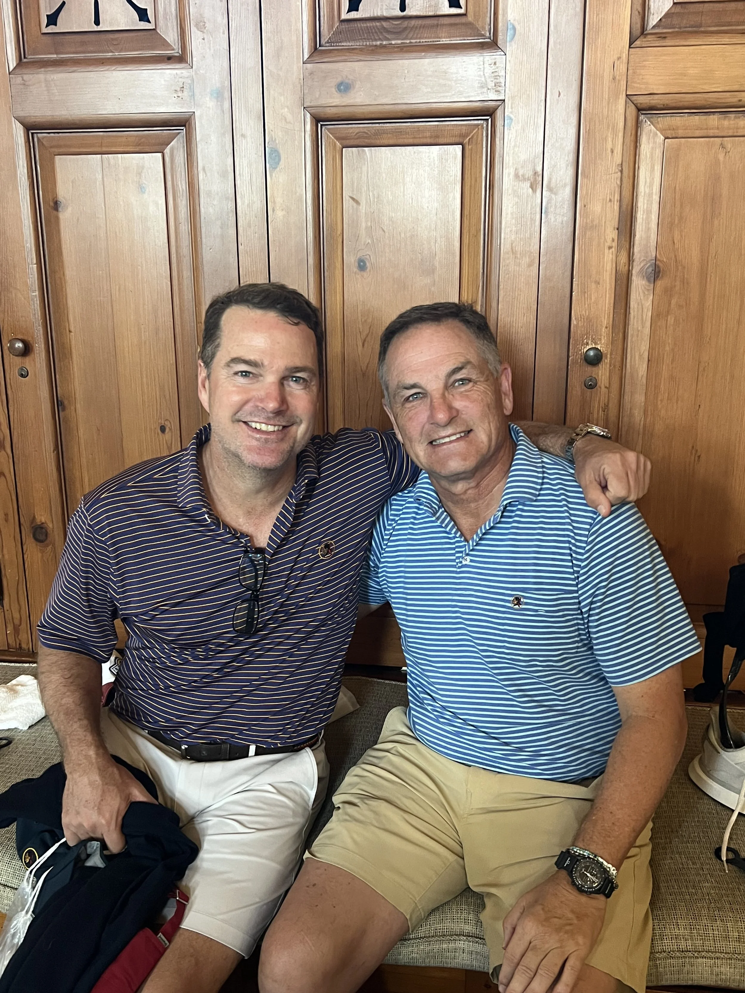 Two men sitting side by side, smiling, with one man having his arm around the other. They are in front of wooden cabinet doors, wearing striped polo shirts and shorts.
