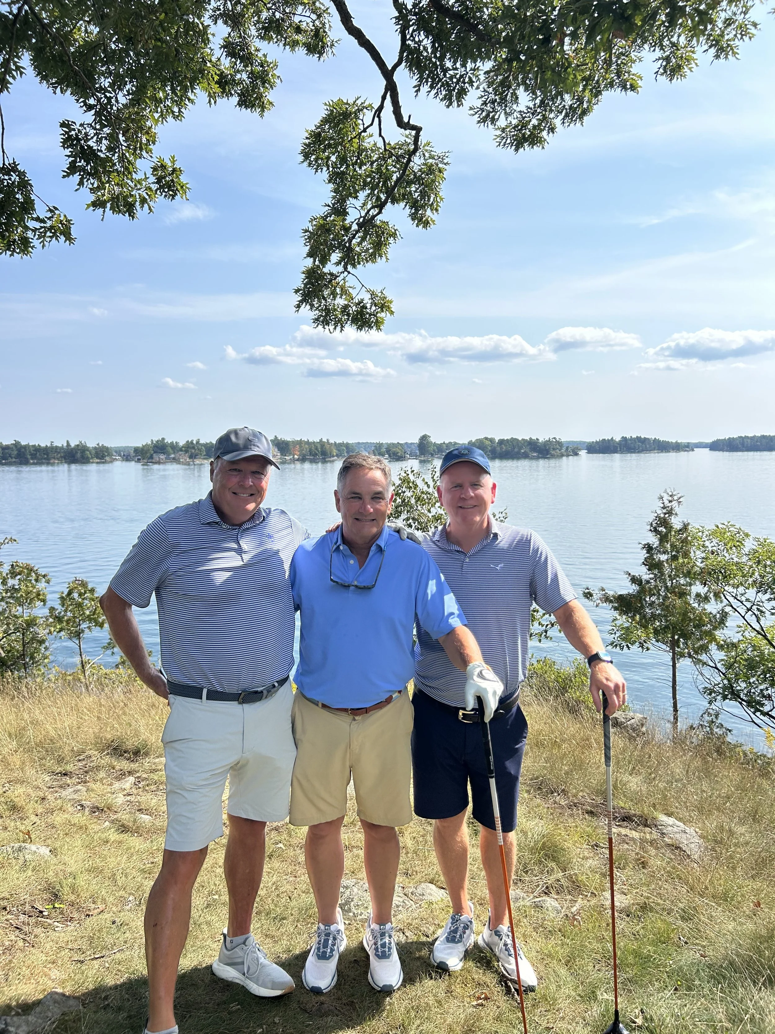 Three men standing together outdoors by the water with trees and a clear blue sky in the background, dressed in golf attire, holding golf clubs.