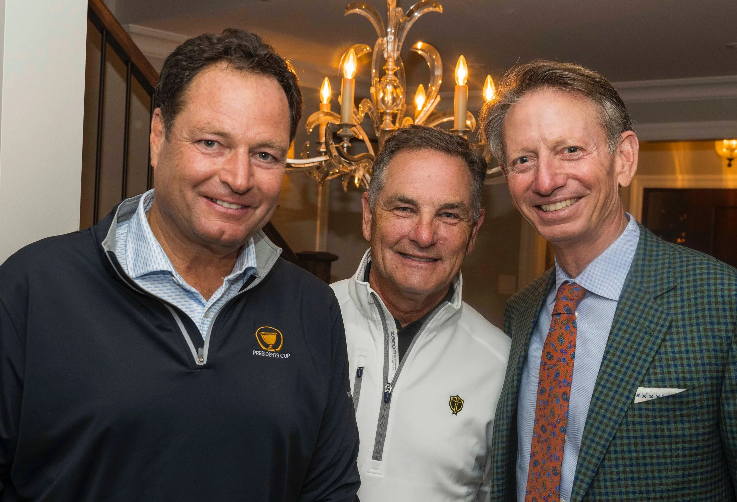 Three men in smart casual attire smiling inside a house with a chandelier overhead.