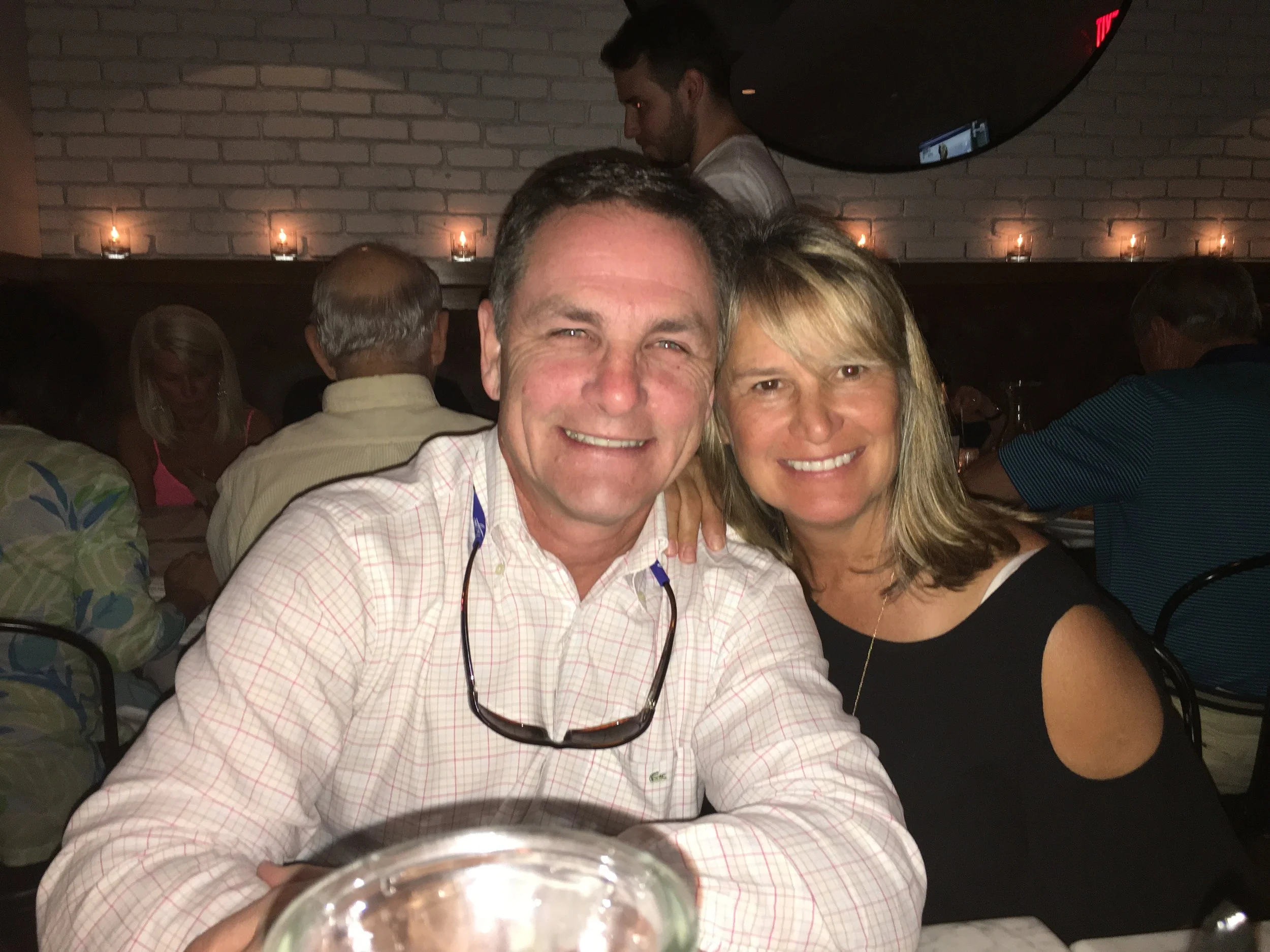 A man and a woman sitting close together at a restaurant, smiling at the camera. The man is wearing a light-colored checkered shirt and sunglasses hanging around his neck, and the woman is wearing a black top with cut-out shoulders. They are surrounded by other diners in a dimly lit setting with candles and a brick wall in the background.