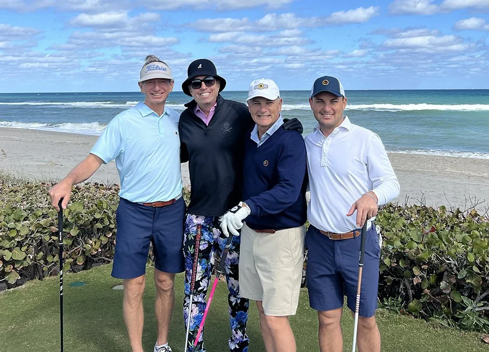 Four men standing together on a golf course near the ocean, holding golf clubs and smiling.