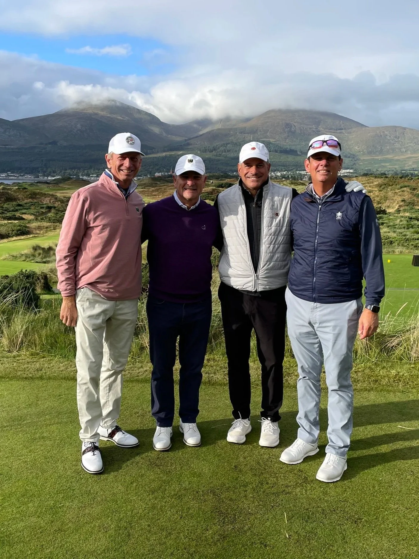 Four men in golf attire standing on a golf course with mountains and cloudy sky in the background.