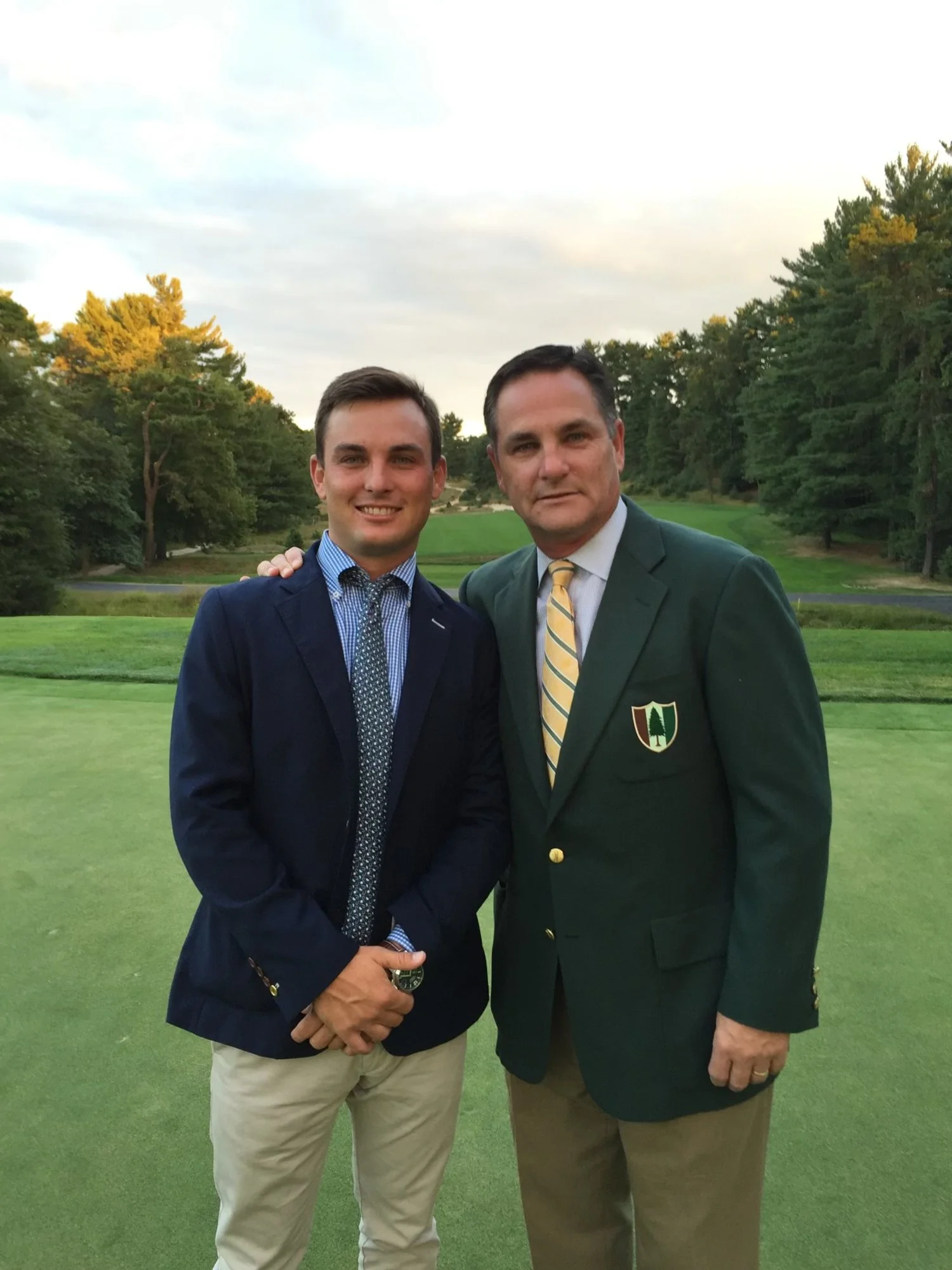 Two men dressed in formal attire standing on a golf course with trees in the background.