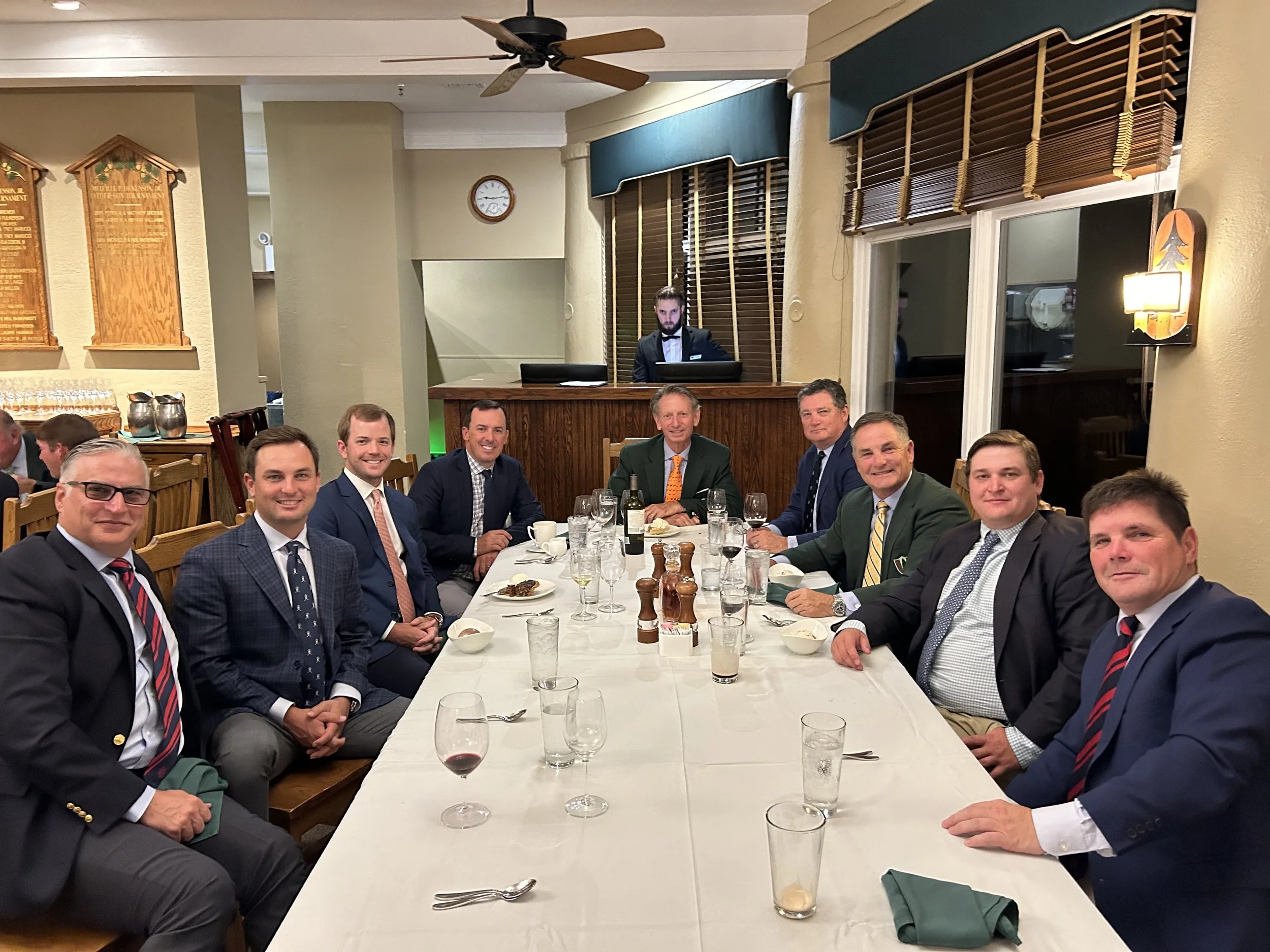 A group of nine men dressed in formal suits seated around a long dining table in a restaurant or banquet hall set for a meal, with some glasses of wine and other drinks on the table, and a background featuring a wooden bar, wall decorations, and a waiter or staff member standing behind the bar.