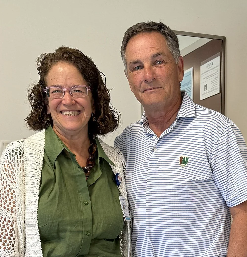A woman with curly hair and glasses smiling next to a man with short hair, both standing indoors in front of a beige wall and a framed notice.