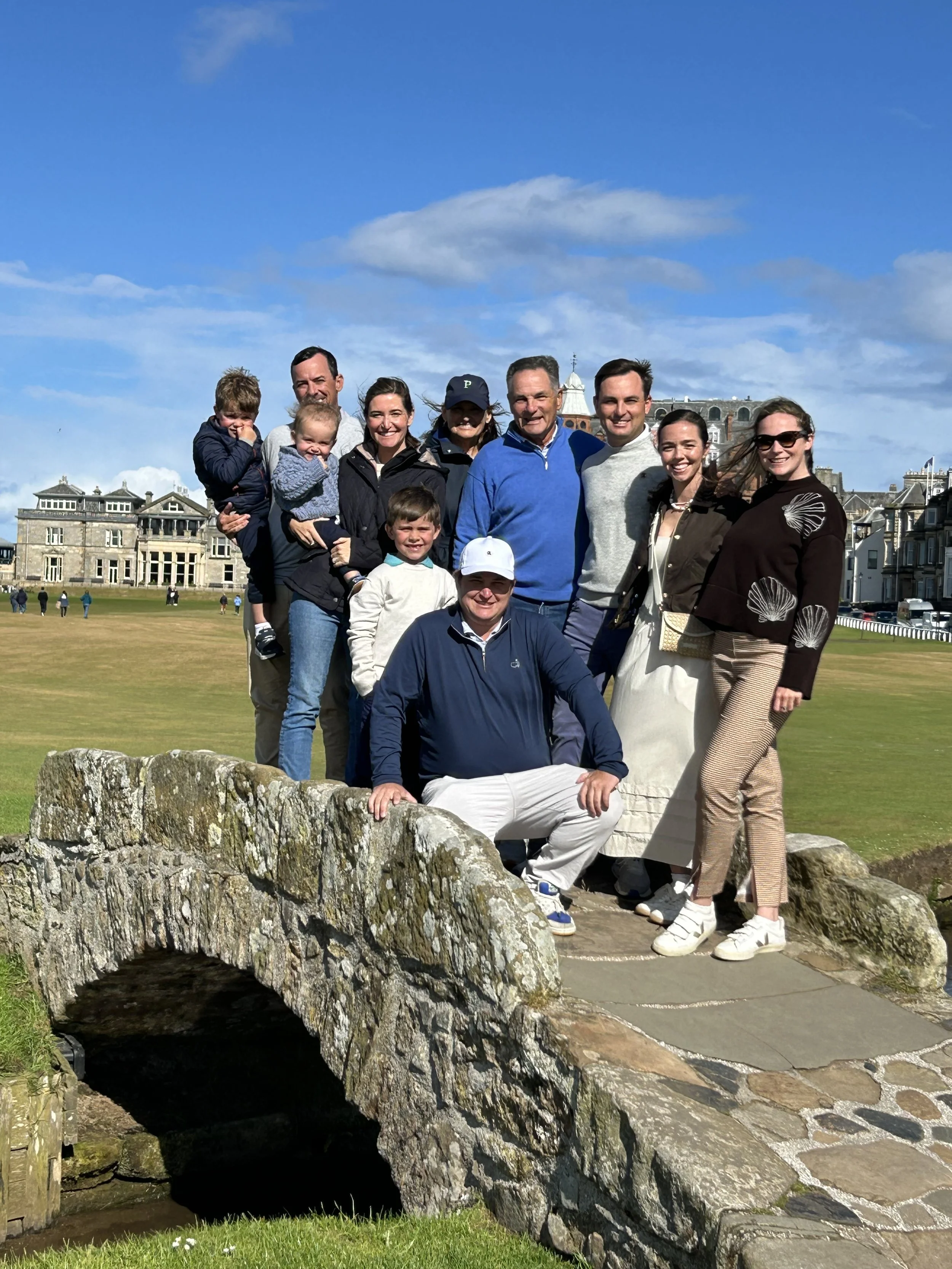 A group of ten people, including children, adults, and seniors, standing on a small stone bridge in a park or field with historic buildings in the background under a blue sky with some clouds.