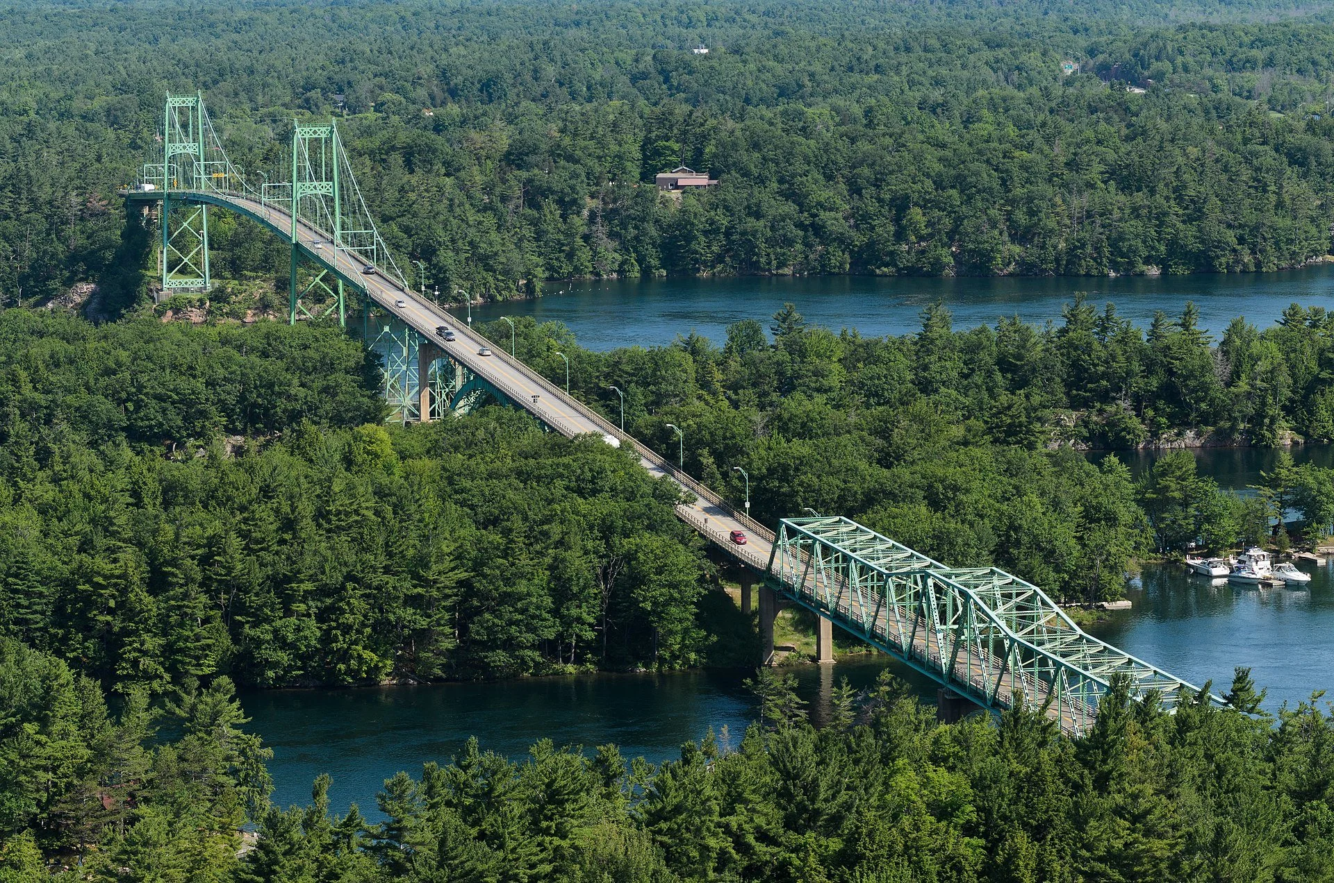Tall green truss bridge crossing over a green forested river with several boats docked along the shoreline, surrounded by dense green trees and hills.