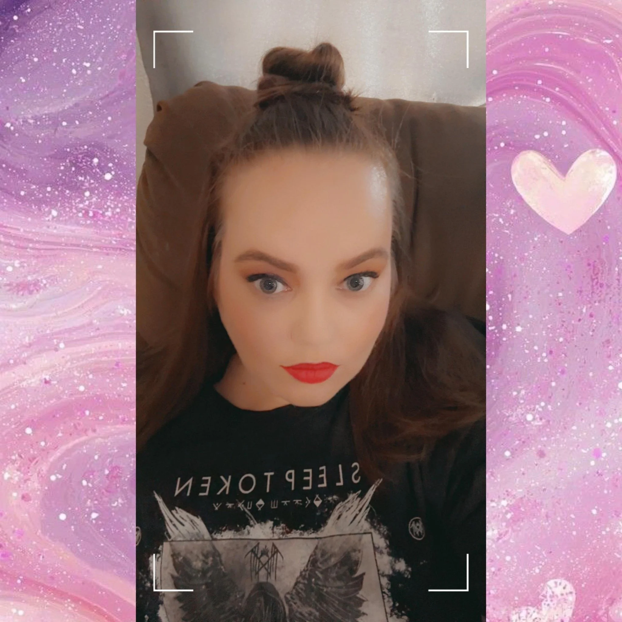 A woman with long red hair styled in a bun, wearing bold red lipstick, a black t-shirt with metal band artwork, sitting on a brown couch, framed by a pink and purple abstract background with a heart illustration.