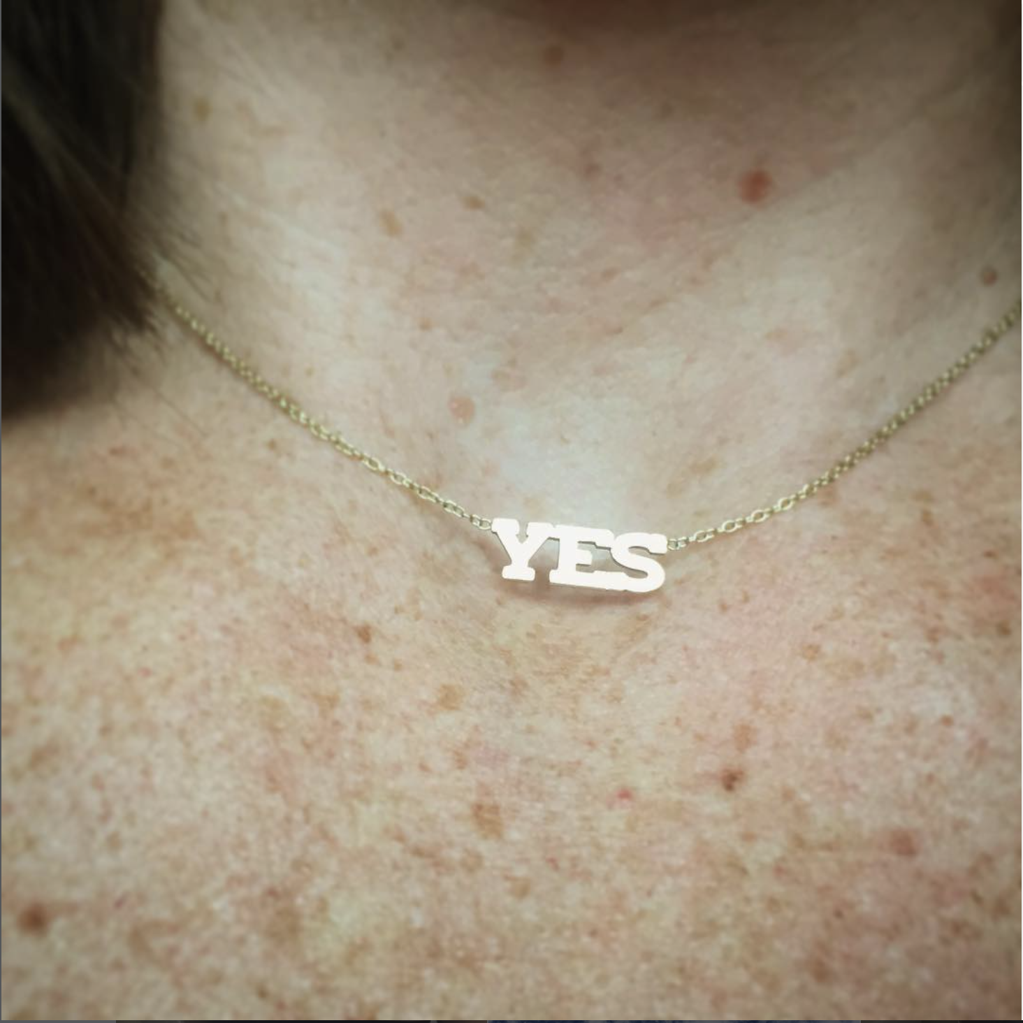 A close-up of a gold necklace with the word 'YES' in capital letters hanging from a chain around a white person's freckled neck.
