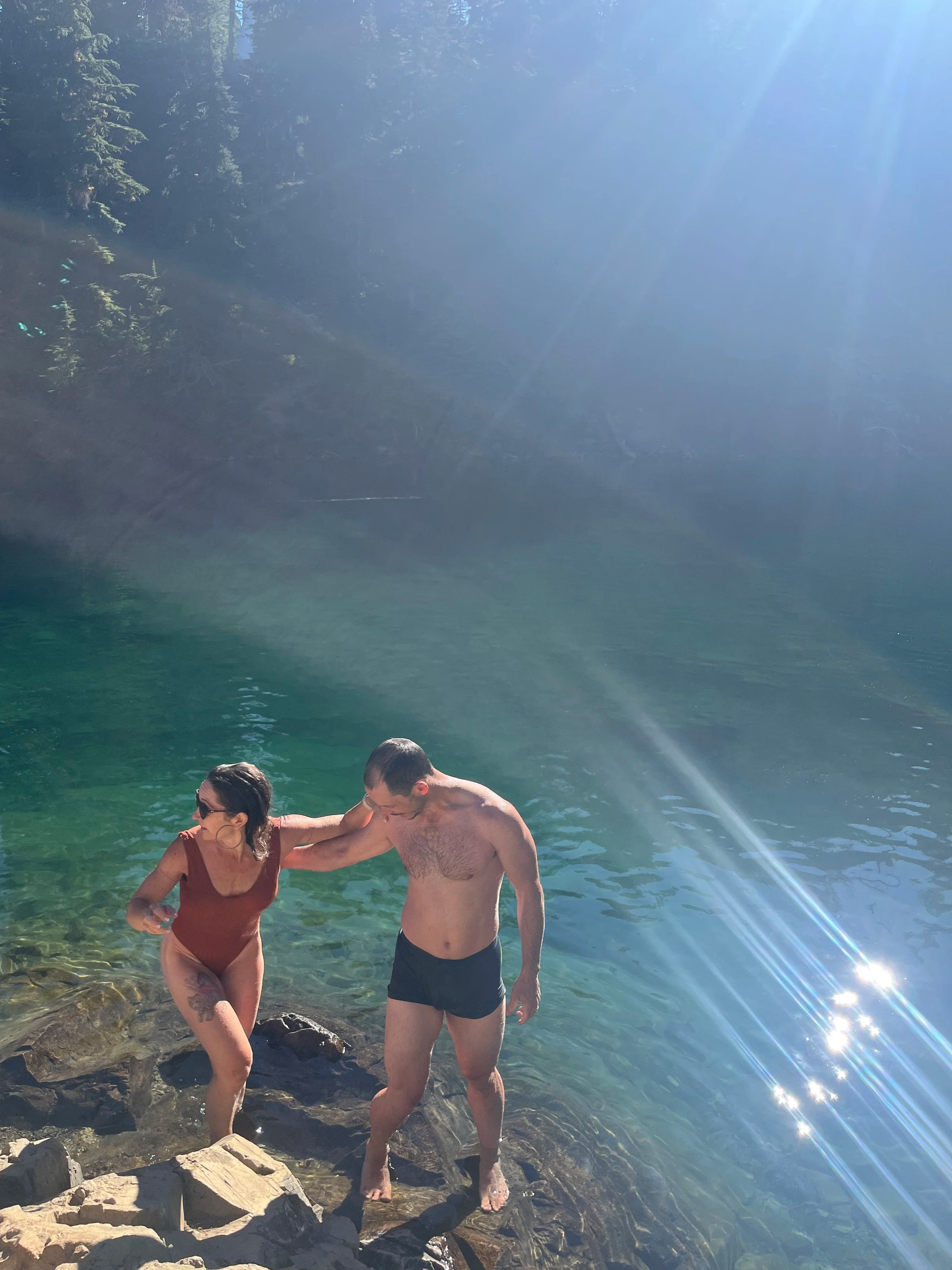A couple is walking out of a body of water, holding hands. The woman is wearing a brown swimsuit, and the man is shirtless wearing black shorts. They are on rocky terrain with clear water and trees in the background, under bright sunlight.