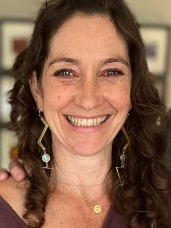 A woman with curly brown hair smiling at the camera, wearing gold earrings and a small gold necklace.