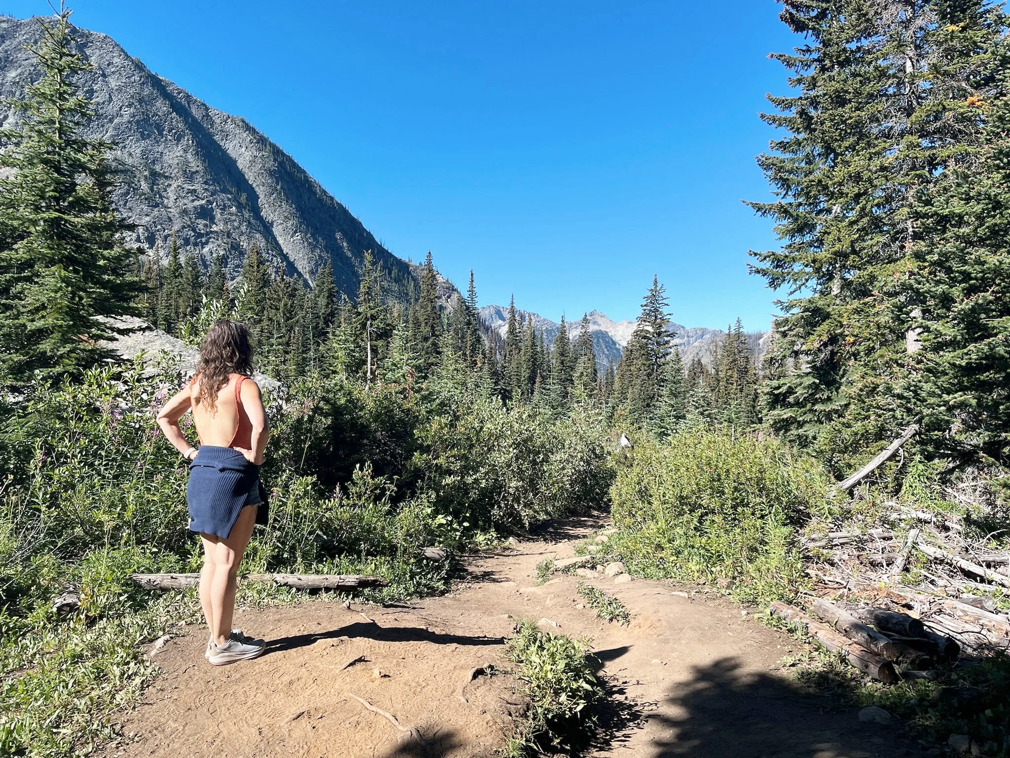 A woman with curly hair and wearing a red top and blue shorts, standing on a dirt trail in a forested mountain landscape in the pacific northwest with tall pine trees, rocky mountain peaks, and a clear blue sky.