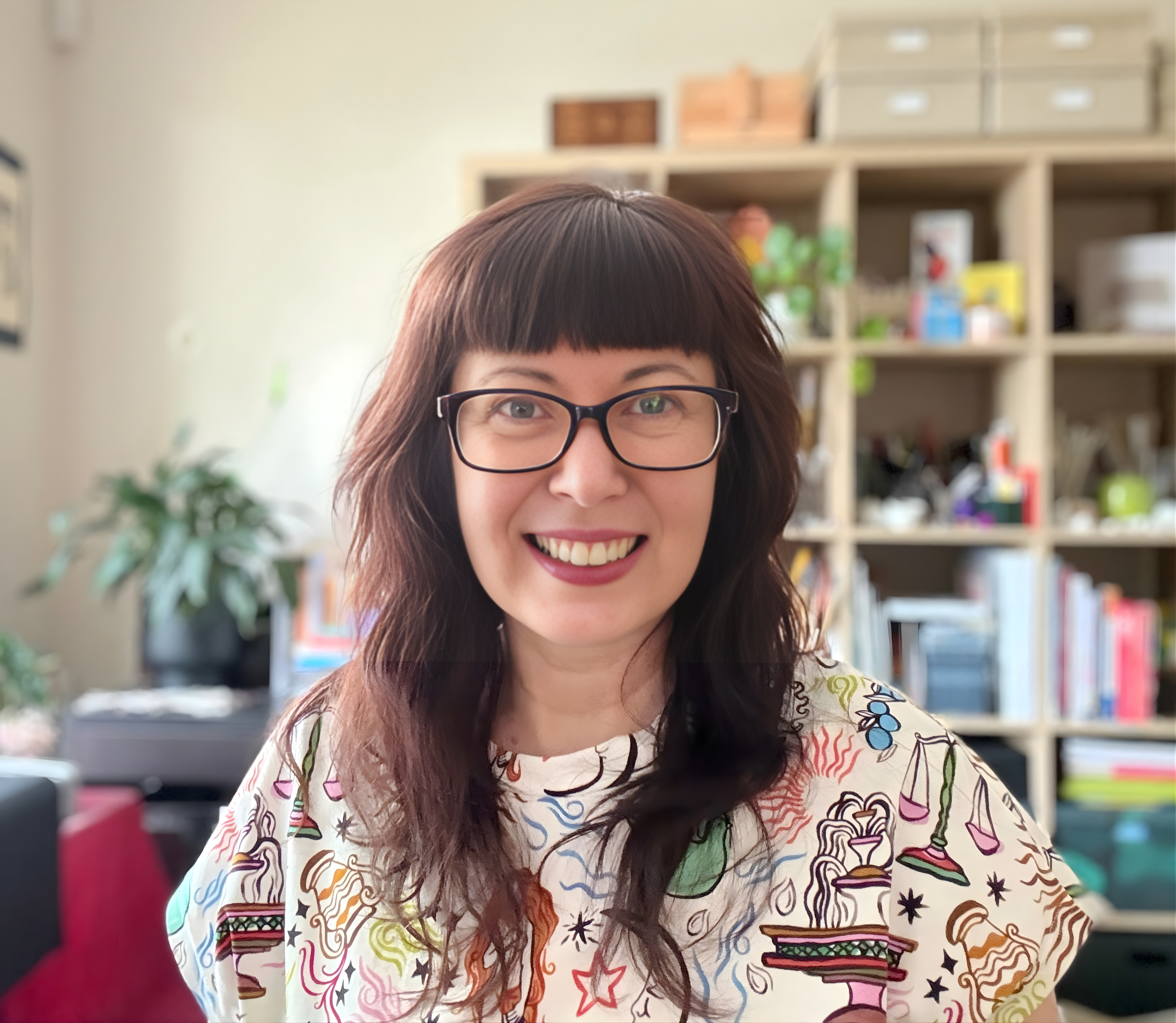 Smiling woman with glasses and brown hair in a colorful shirt, sitting in a room with bookshelves and houseplants in the background.