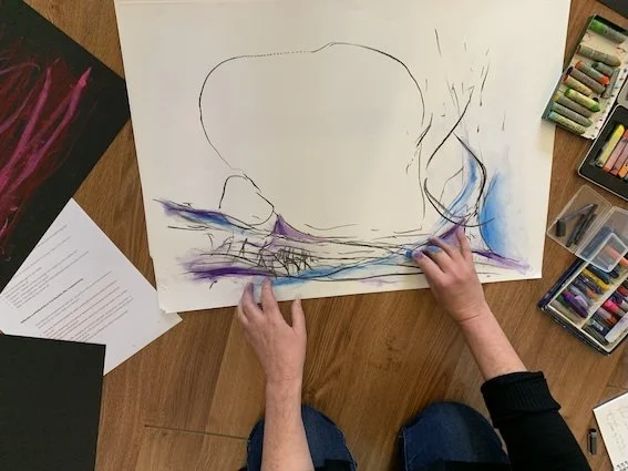 Person sitting on the floor creating abstract artwork with pastels on large paper in a well-lit room.