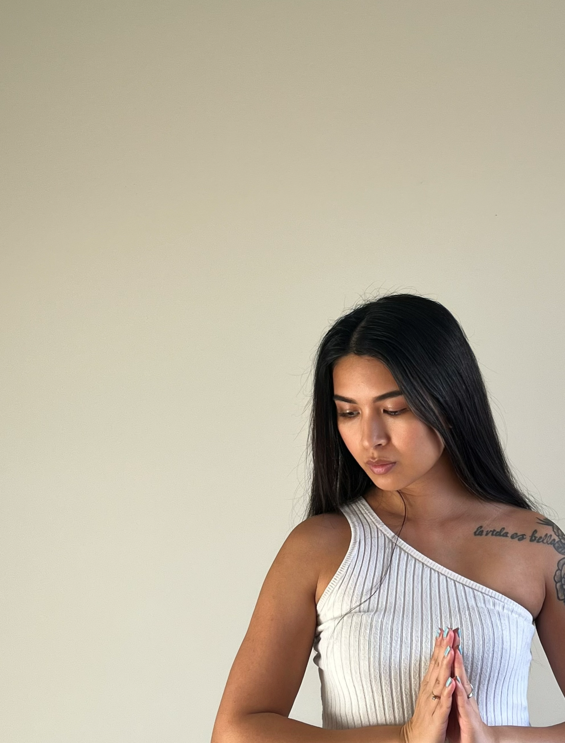 Young woman with long black hair in white sleeveless top, her hands pressed together in prayer, standing against a plain light-colored wall.